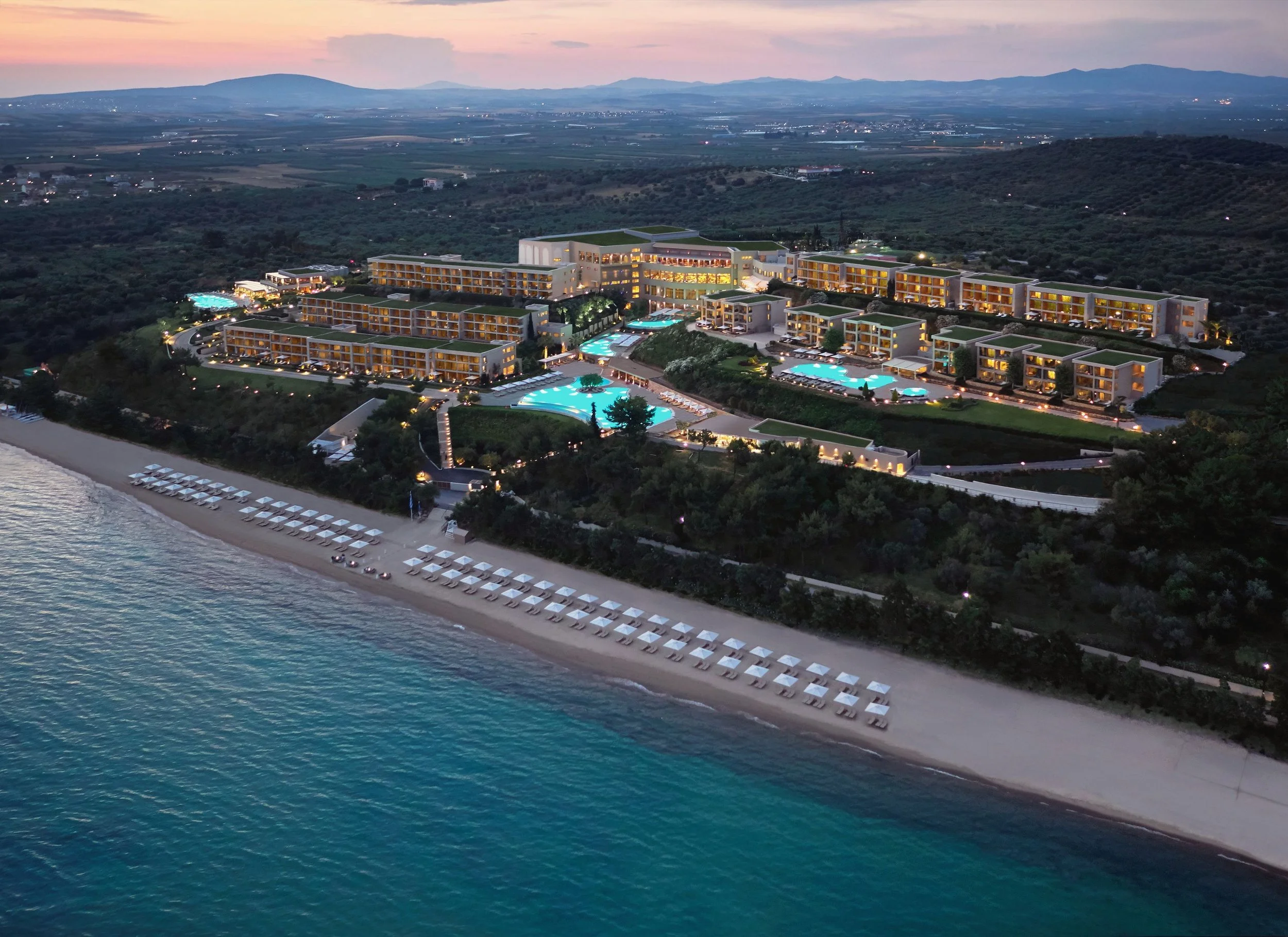 An aerial view of a luxury seaside resort at dusk with multiple swimming pools, beachfront, and modern multi-story buildings surrounded by lush greenery.