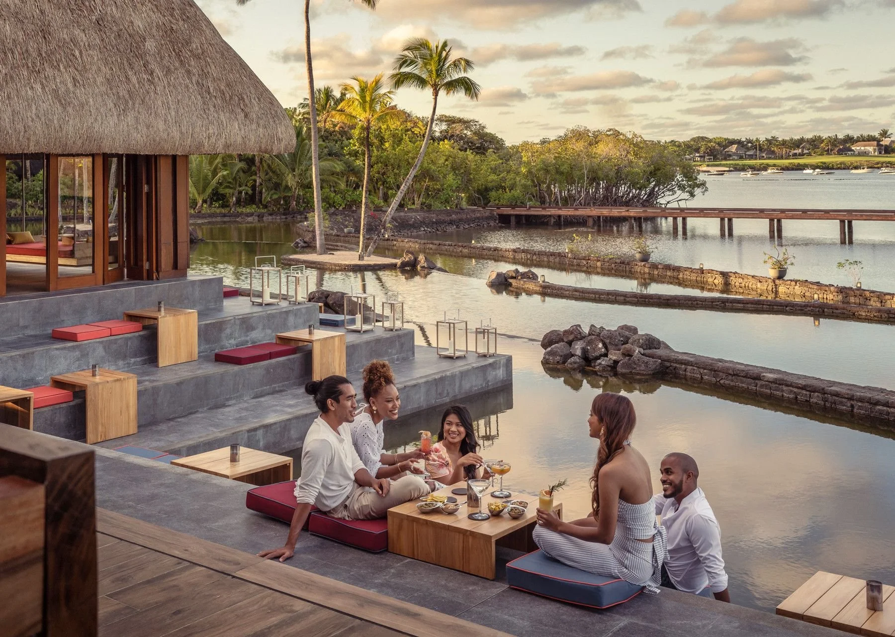 A group of six friends enjoying drinks and conversation on a patio overlooking a calm waterway with boats and lush greenery in the background during sunset at a tropical resort.
