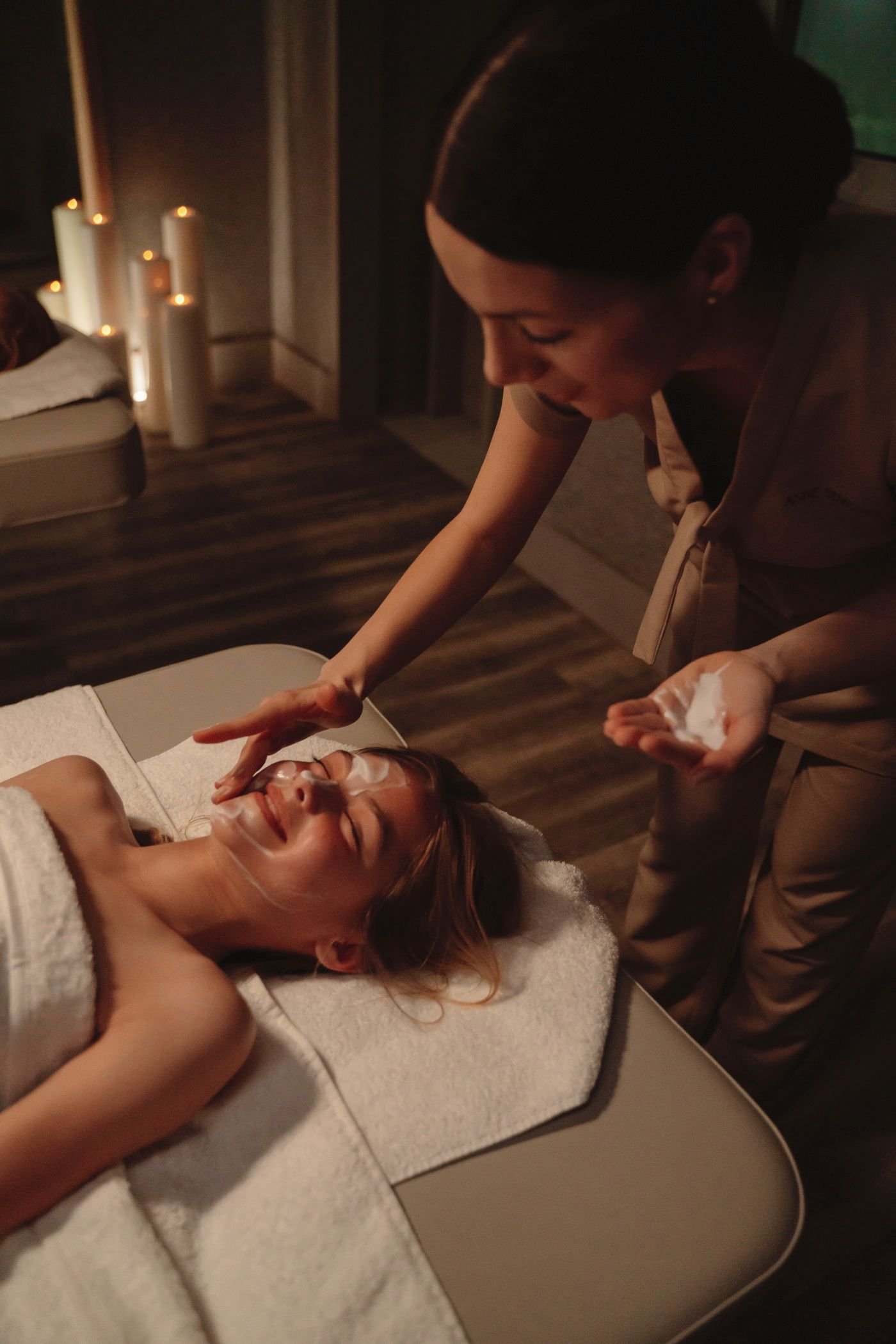 A woman is lying on a massage table covered with a towel, receiving a facial treatment. A professional esthetician is applying a white cream or mask to her face in a dimly lit spa-like setting with candles in the background.