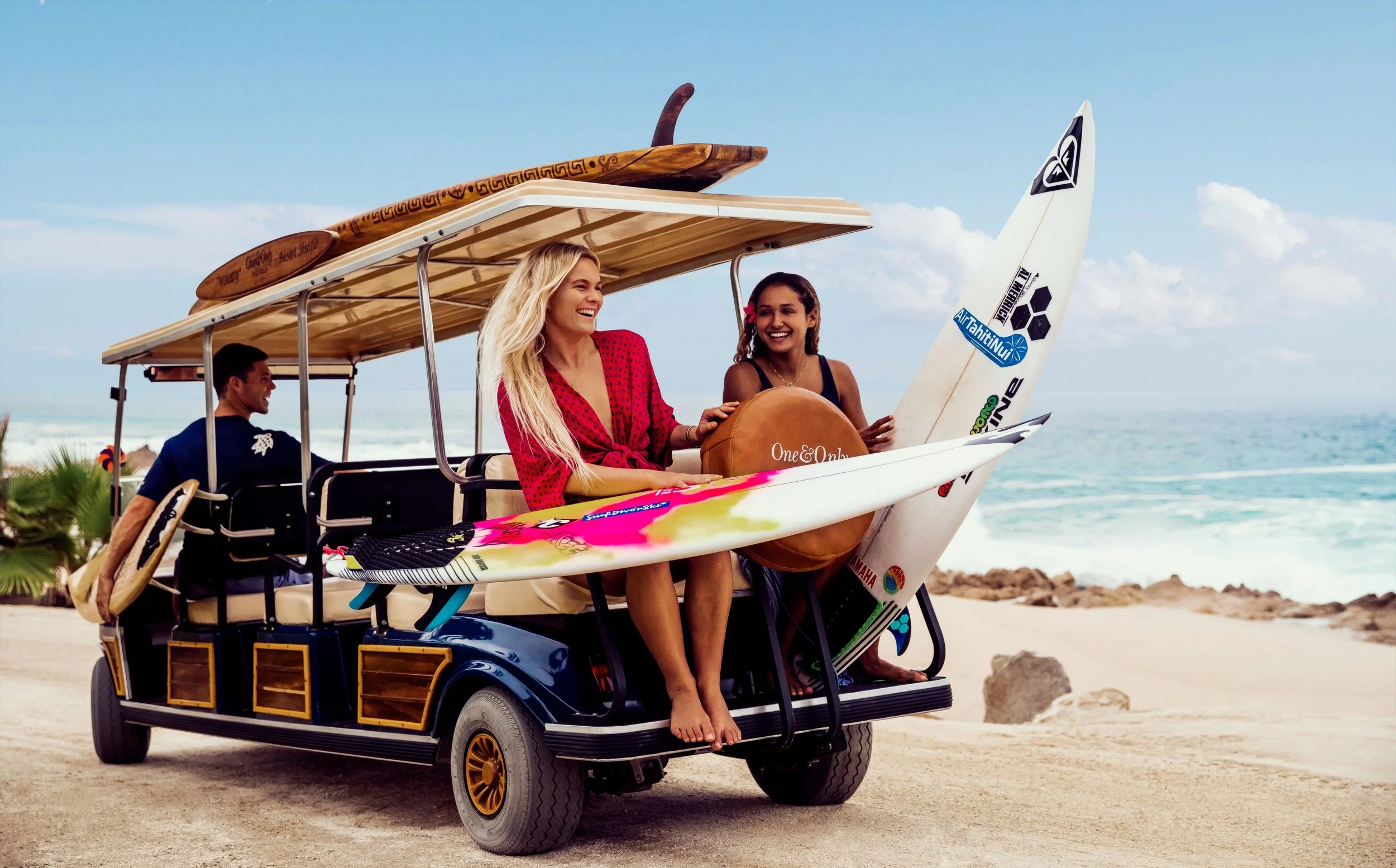 Three women riding in an open-air golf cart on a beach, carrying surfboards, with the ocean and a rocky shoreline in the background.