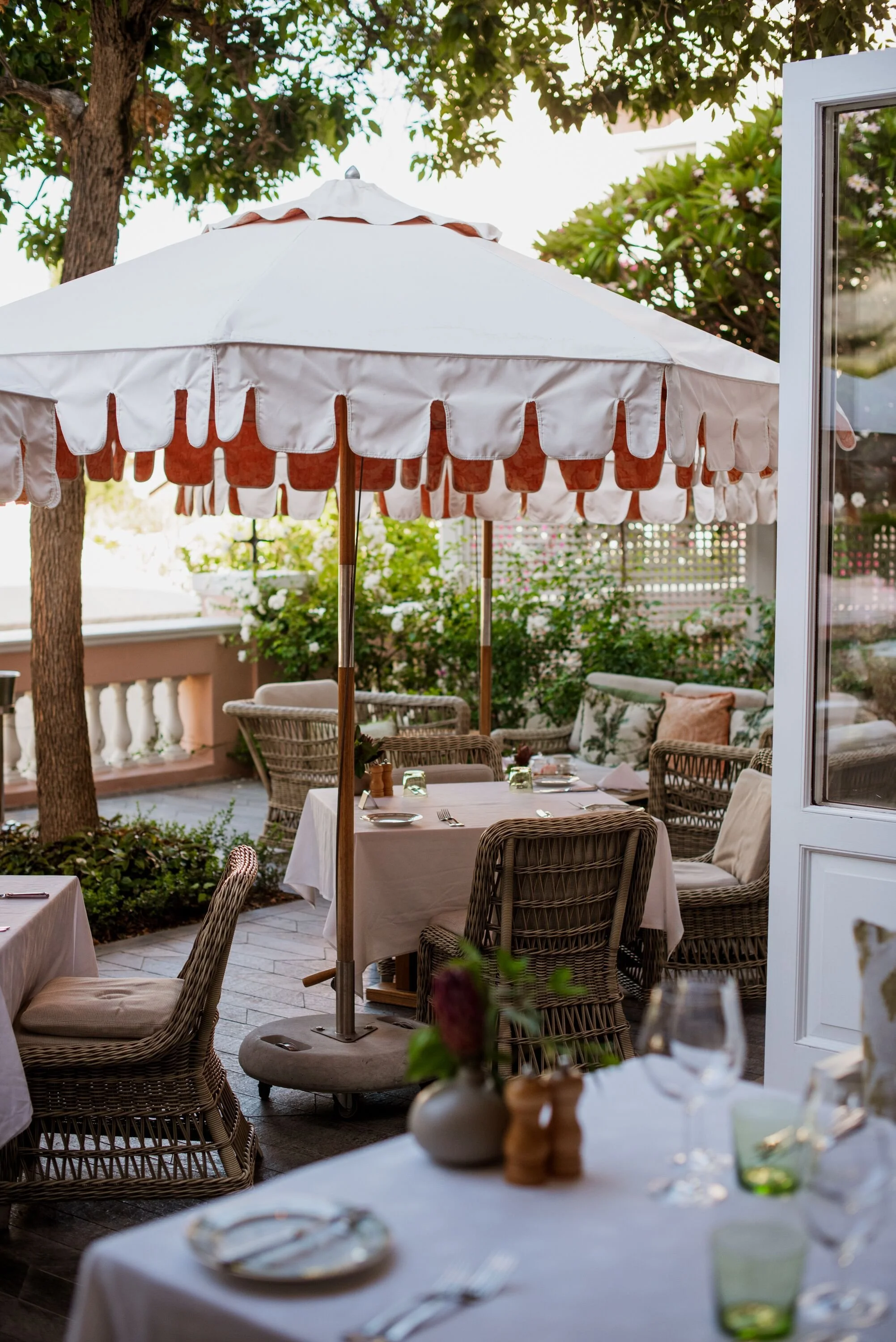 An outdoor dining area with tables covered with white tablecloths, surrounded by wicker chairs. A large white patio umbrella shades the space, with greenery including trees and bushes in the background. The setting appears to be a garden or patio.