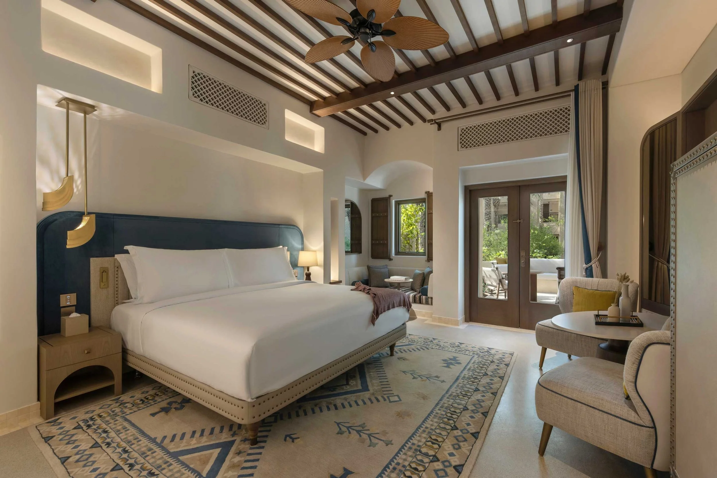 A bedroom with a bed that has a blue headboard, a nightstand with a lamp, and a ceiling fan with leaf-shaped blades. There are windows and a sliding door leading to a balcony or patio, with outdoor furniture visible outside. The room has white walls, wooden accents, and patterned area rugs.