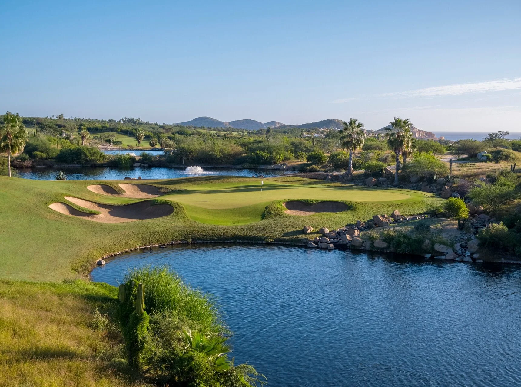 A scenic view of a golf course with a green, sand bunkers, and water hazards, surrounded by desert plants and palm trees, with a mountainous landscape in the background under a clear blue sky.
