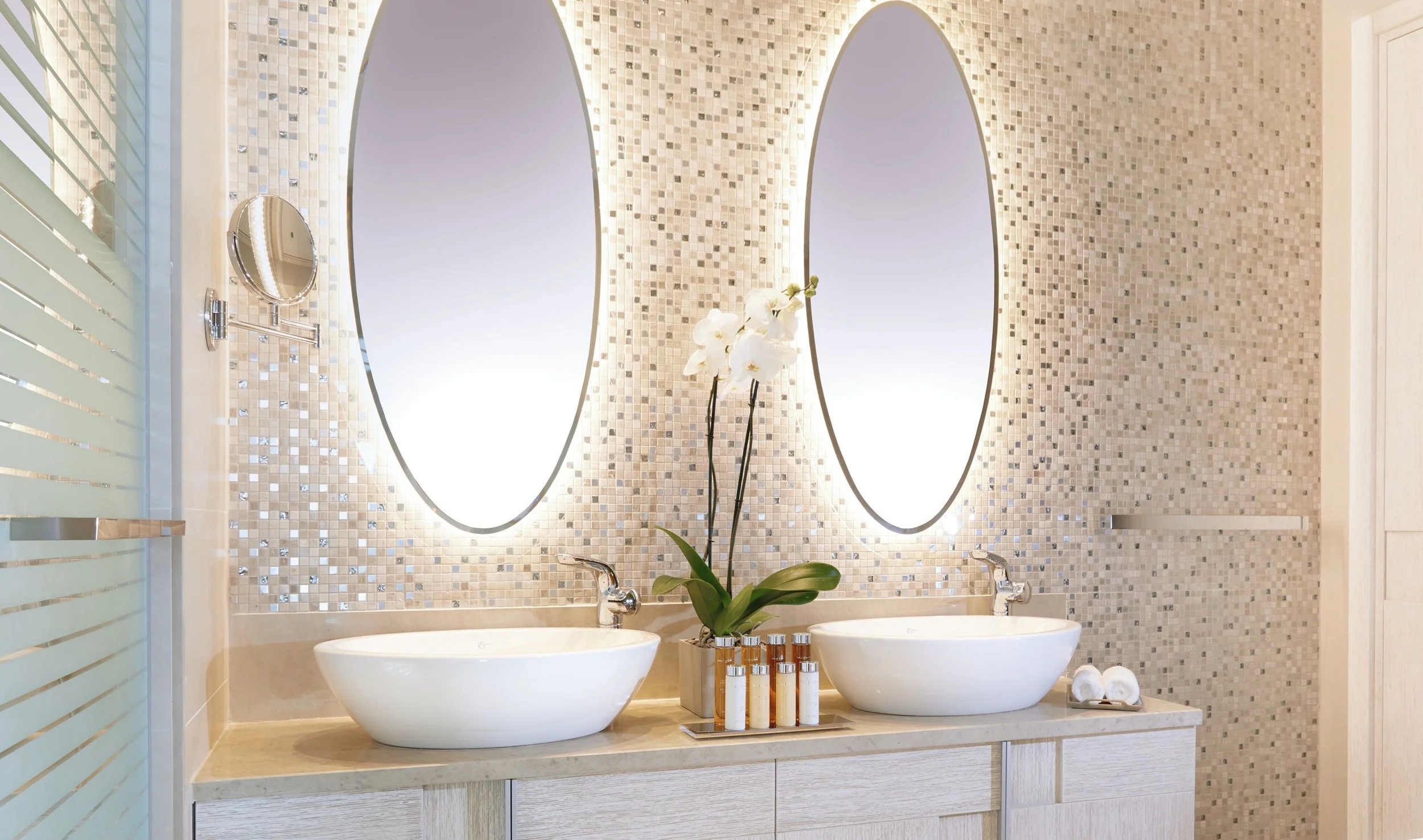 A modern bathroom vanity with two vessel sinks, a large mirror, and a small potted orchid plant in the center. The wall behind features small mosaic tiles, and there's lighting around the mirror.