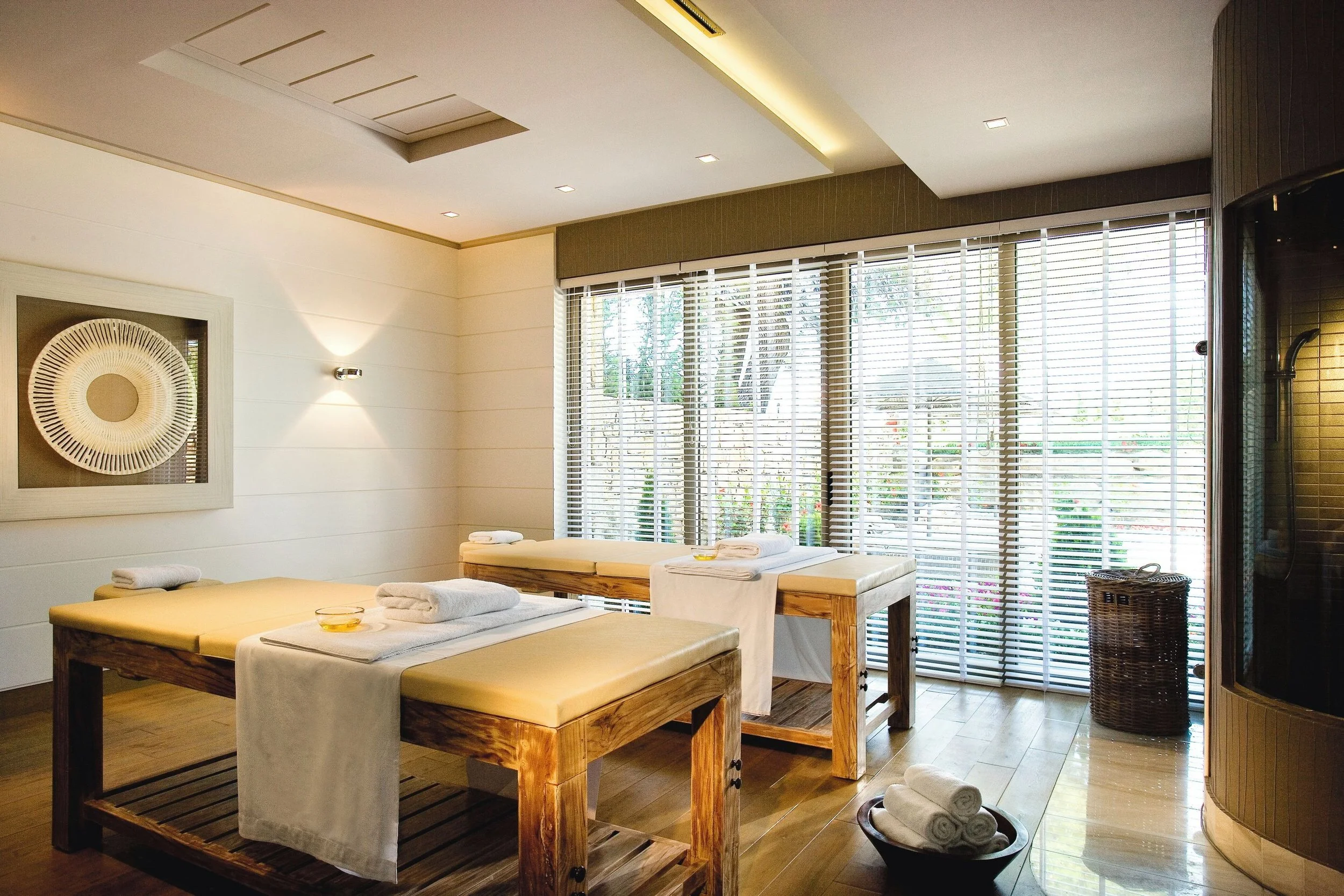 Spa or massage room with two massage tables, rolled towels, and large windows with blinds, sunlit and overlooking trees.