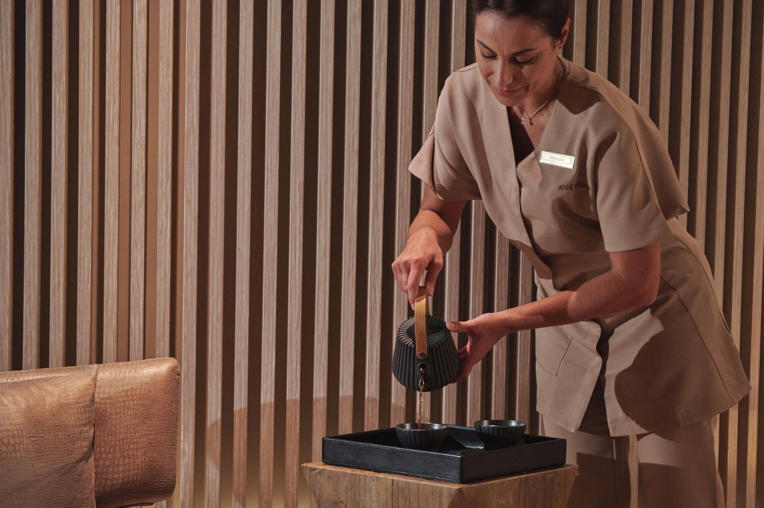 A woman in a beige uniform with a name tag that reads Olympia, pouring liquid from a metal teapot into small black bowls on a wooden tray.