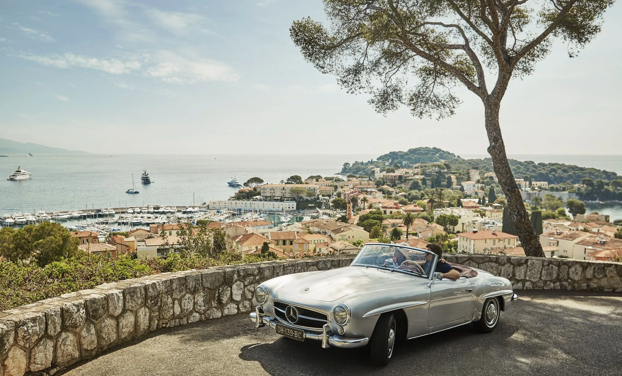 A vintage silver Mercedes-Benz convertible car parked next to a stone wall overlooking a coastal town with houses, trees, a marina with boats, and a large tree with a thick trunk and spreading branches in the background.