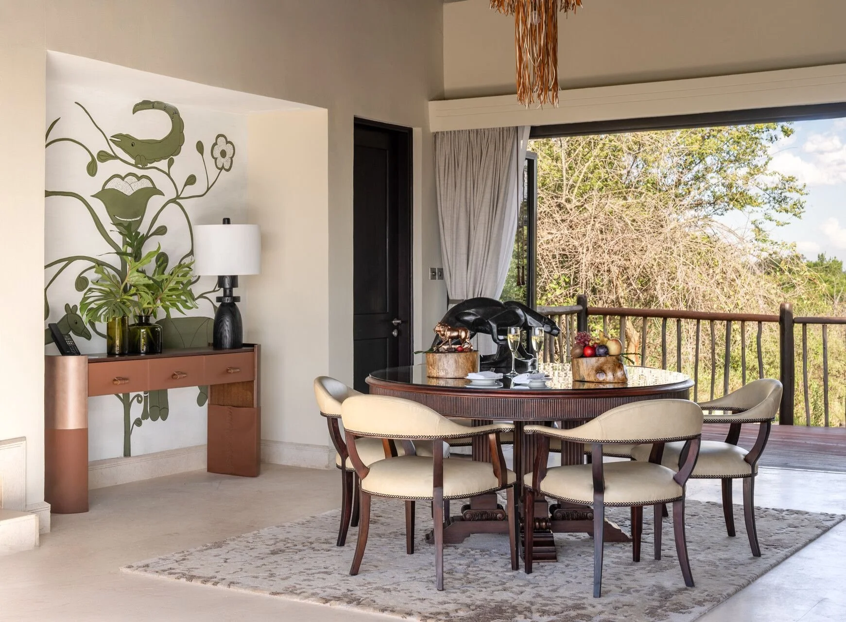A dining room with a round wooden table surrounded by six beige upholstered chairs. The table has a sculpture, a bowl of fruit, and a glass of champagne. There is a large window with open curtains revealing trees outside. To the left, a sideboard with a black lamp, a telephone, and decorative items is against a wall with a green and white floral and animal mural.