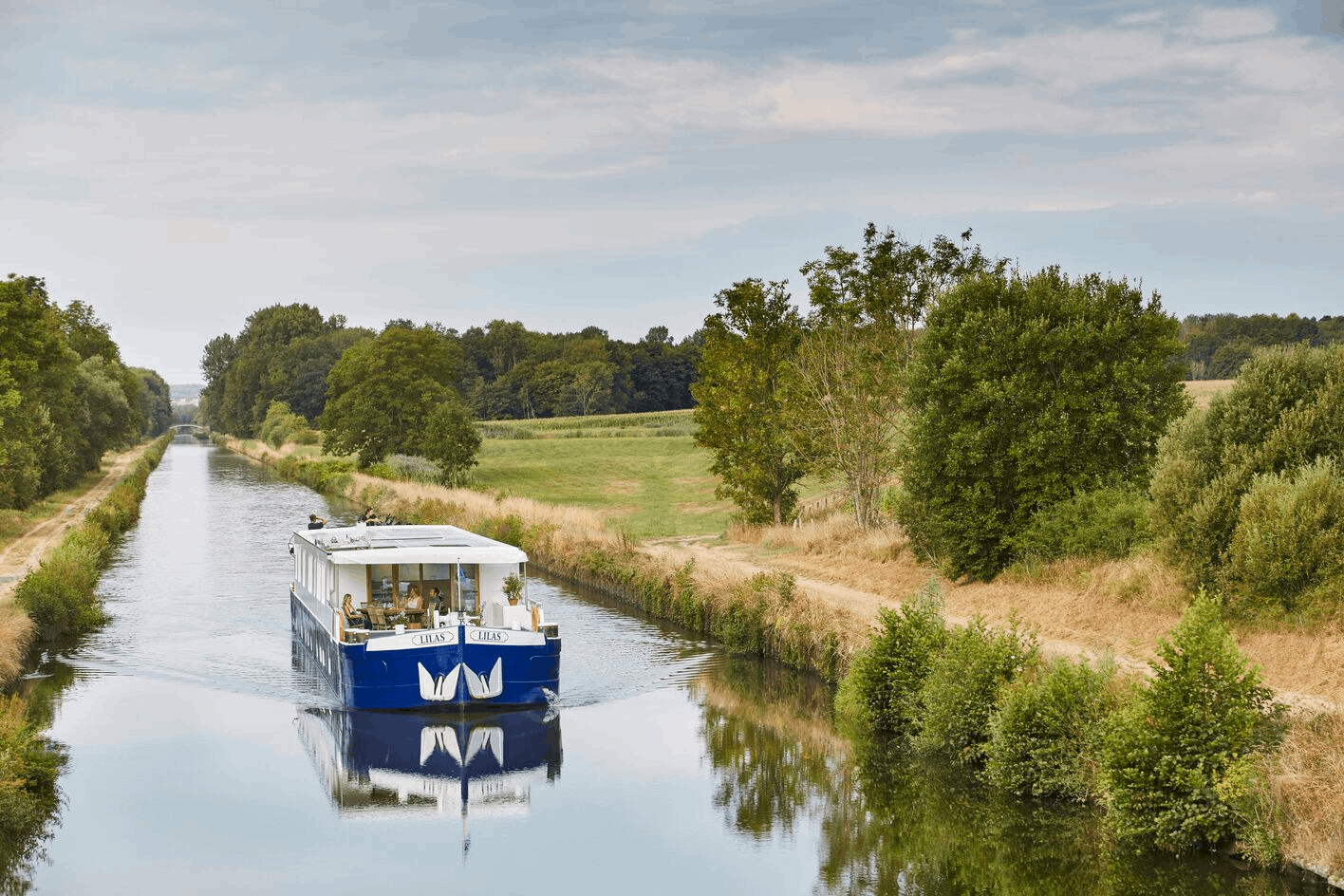 A blue and white boat named 'TILAS' sailing on a calm, narrow river surrounded by green trees and grassy fields under a blue sky with clouds.