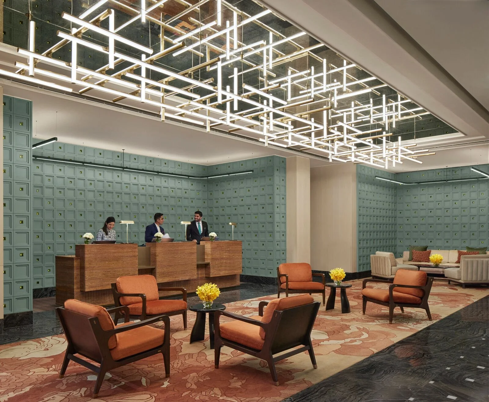 A hotel lobby with a reception desk, three staff members behind it, and seating area with peach and beige chairs and sofas, decorated with yellow flowers, under a modern geometric light fixture.
