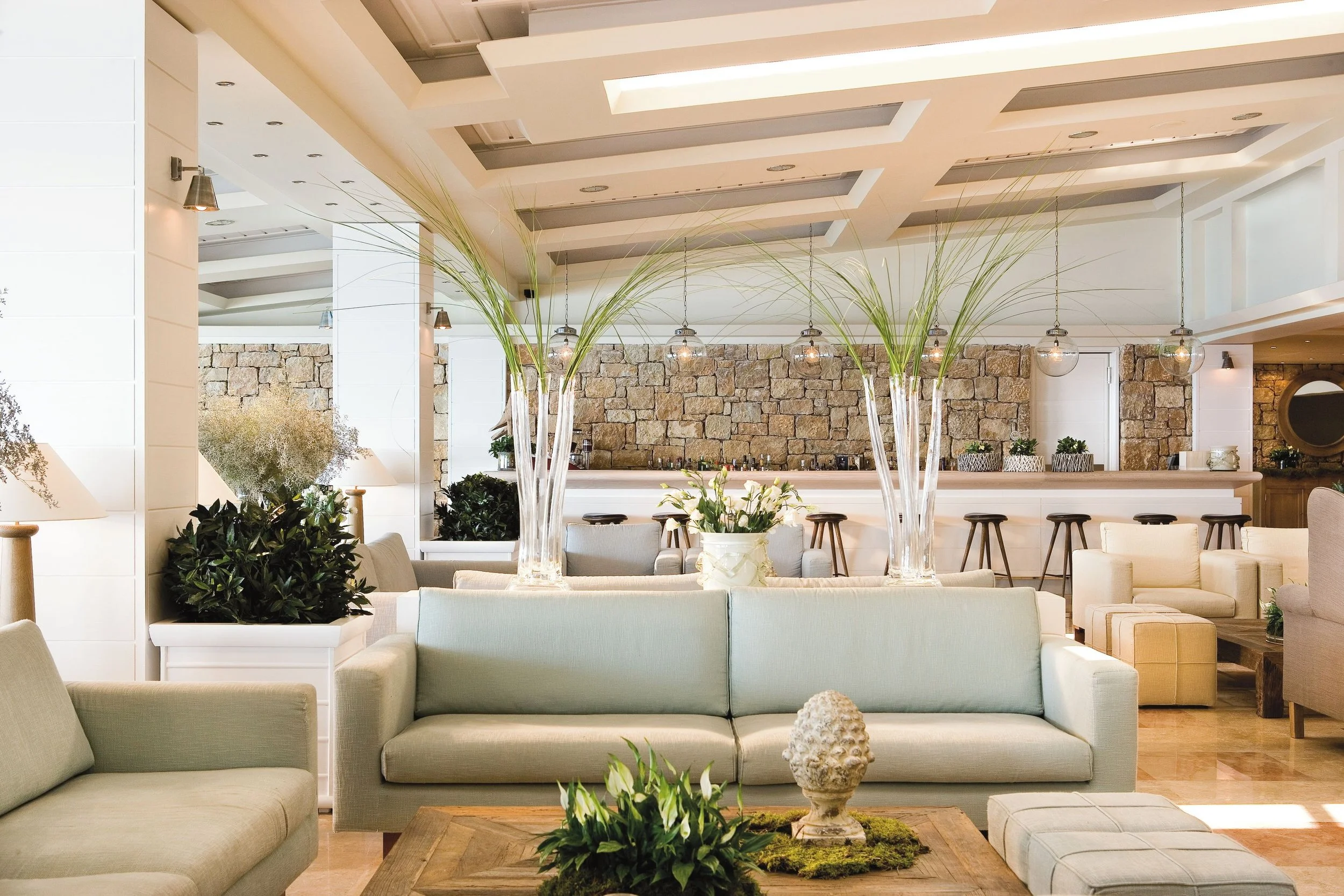 Modern interior of a lounge or hotel lobby with beige sofas, potted plants, a stone accent wall, and decorative lighting fixtures hanging from the ceiling.
