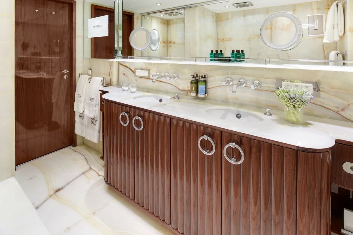 A hotel bathroom with a double sink vanity, wooden cabinetry, marble walls, and a large mirror.