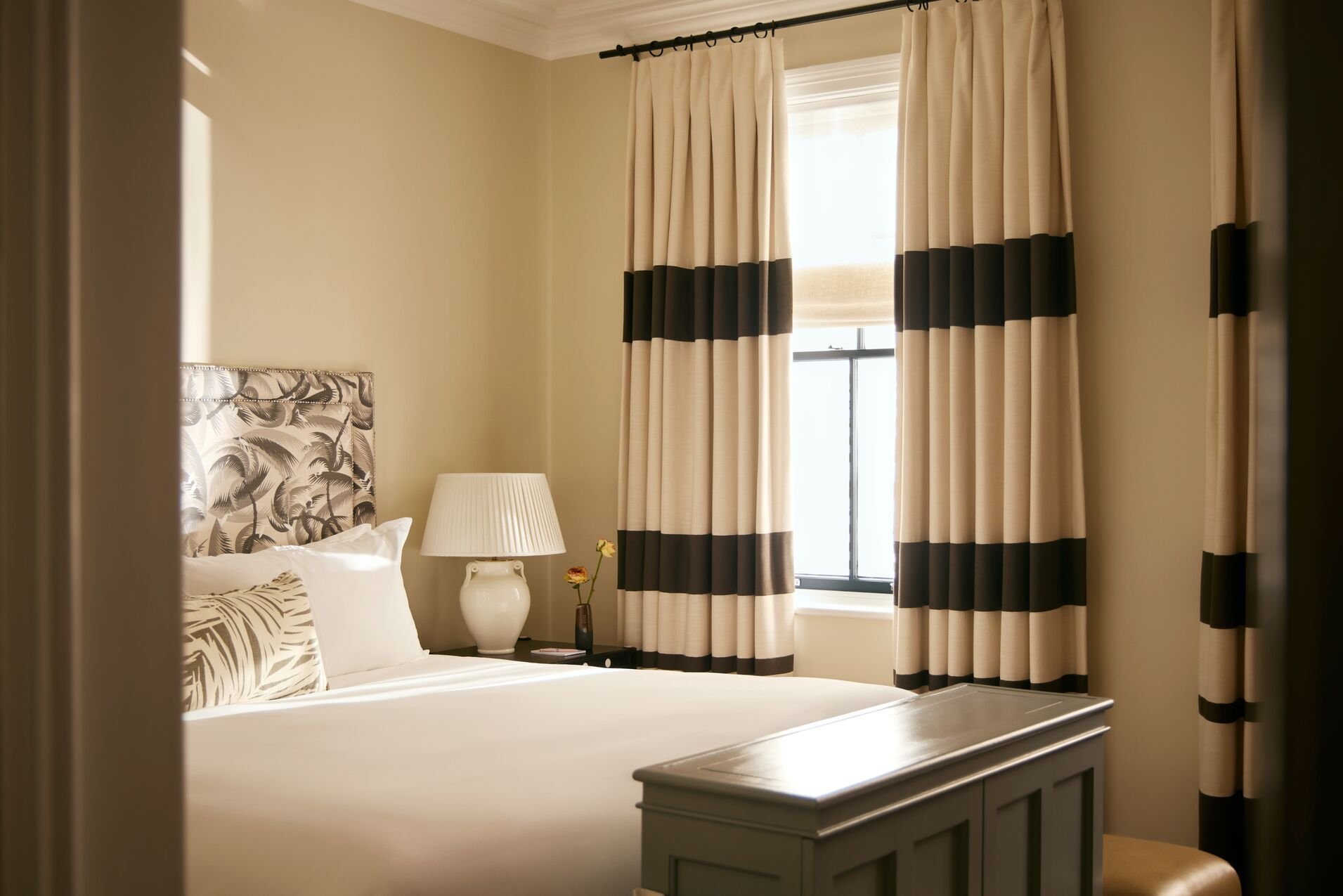 A cozy bedroom with a neatly made bed, patterned headboard, white pillows, a nightstand with a white lamp, a small vase with flowers, and large cream-colored curtains with black stripes covering the window.