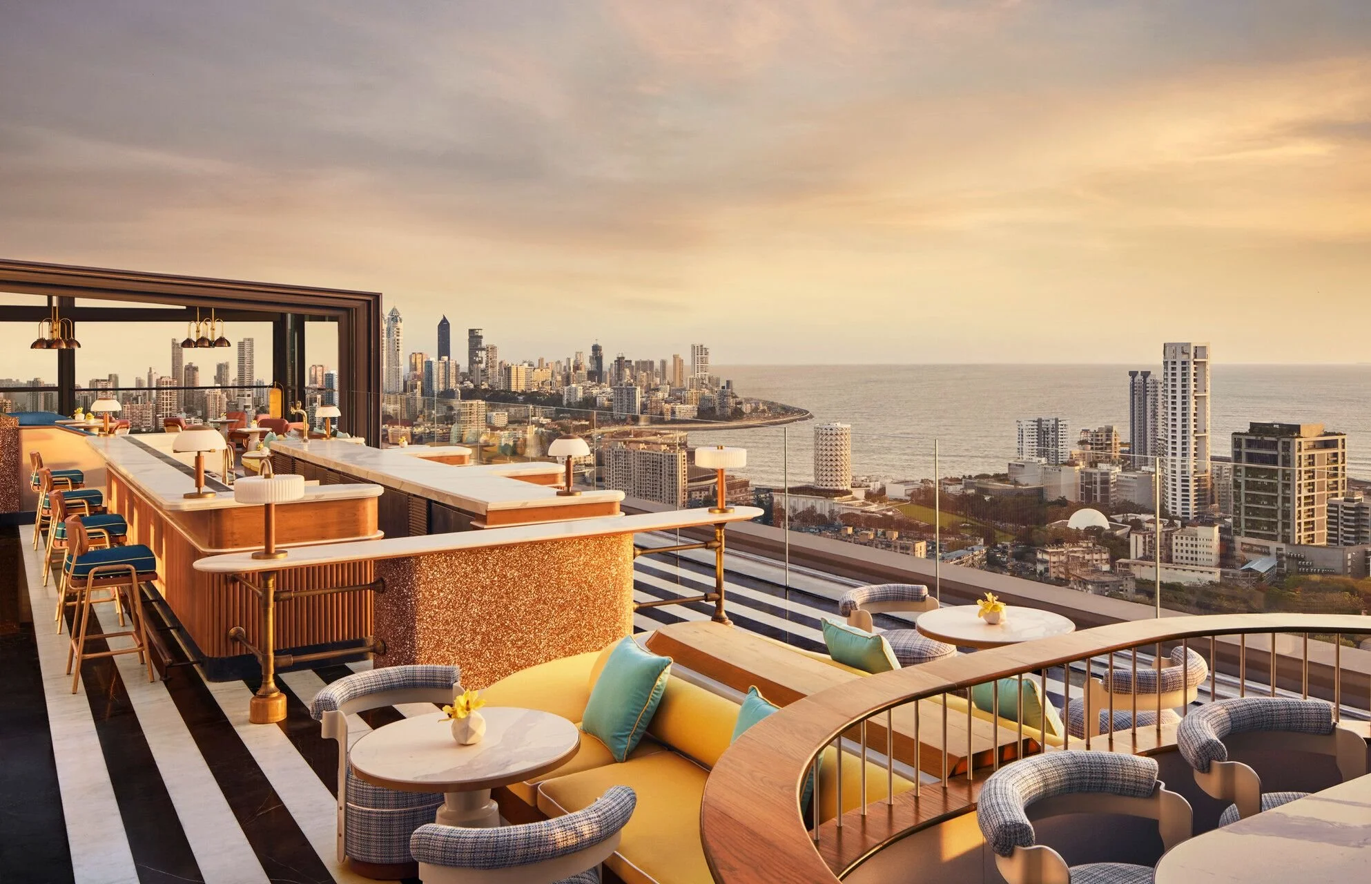 Rooftop bar with city skyline and ocean view, modern furniture, tables with small flower arrangements, and a sunset sky.