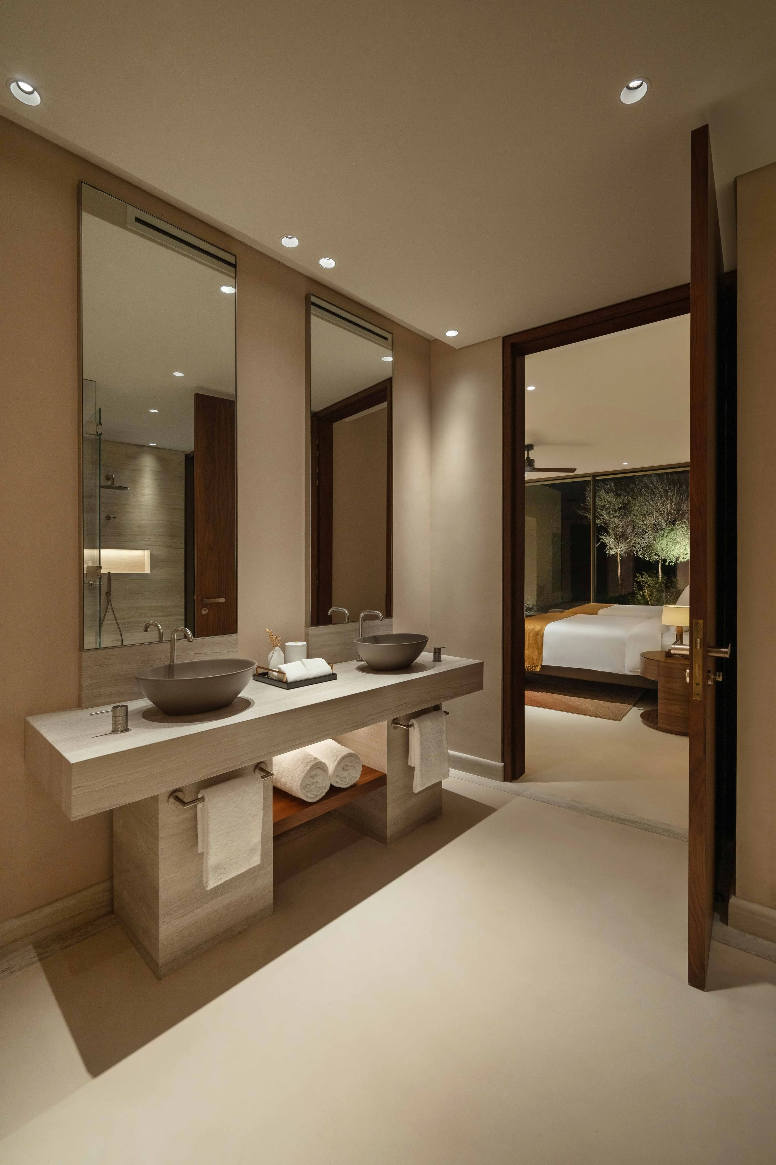 Modern bathroom with two vessel sinks on a wooden vanity, large wall mirrors, and a view into a bedroom with a large bed and sliding glass door leading outside.