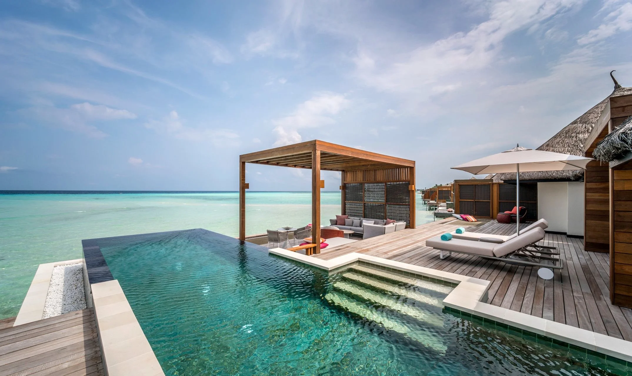 Luxury overwater bungalow with infinity pool, lounge chairs, cabana, and ocean view under cloudy sky.
