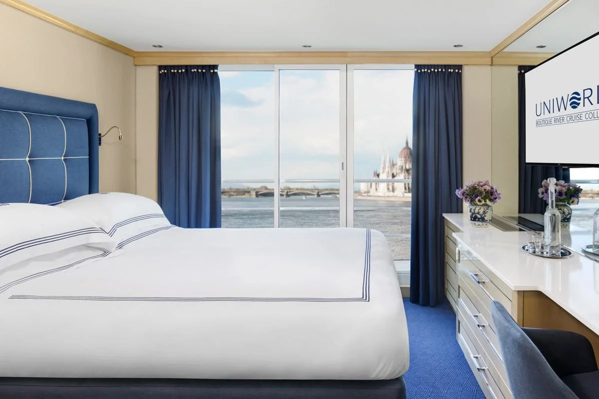 Interior of a luxury hotel room with a large bed, blue headboard, white bedding, and a view of a river and historical building through large windows, with blue curtains and a white desk with flowers and a TV.