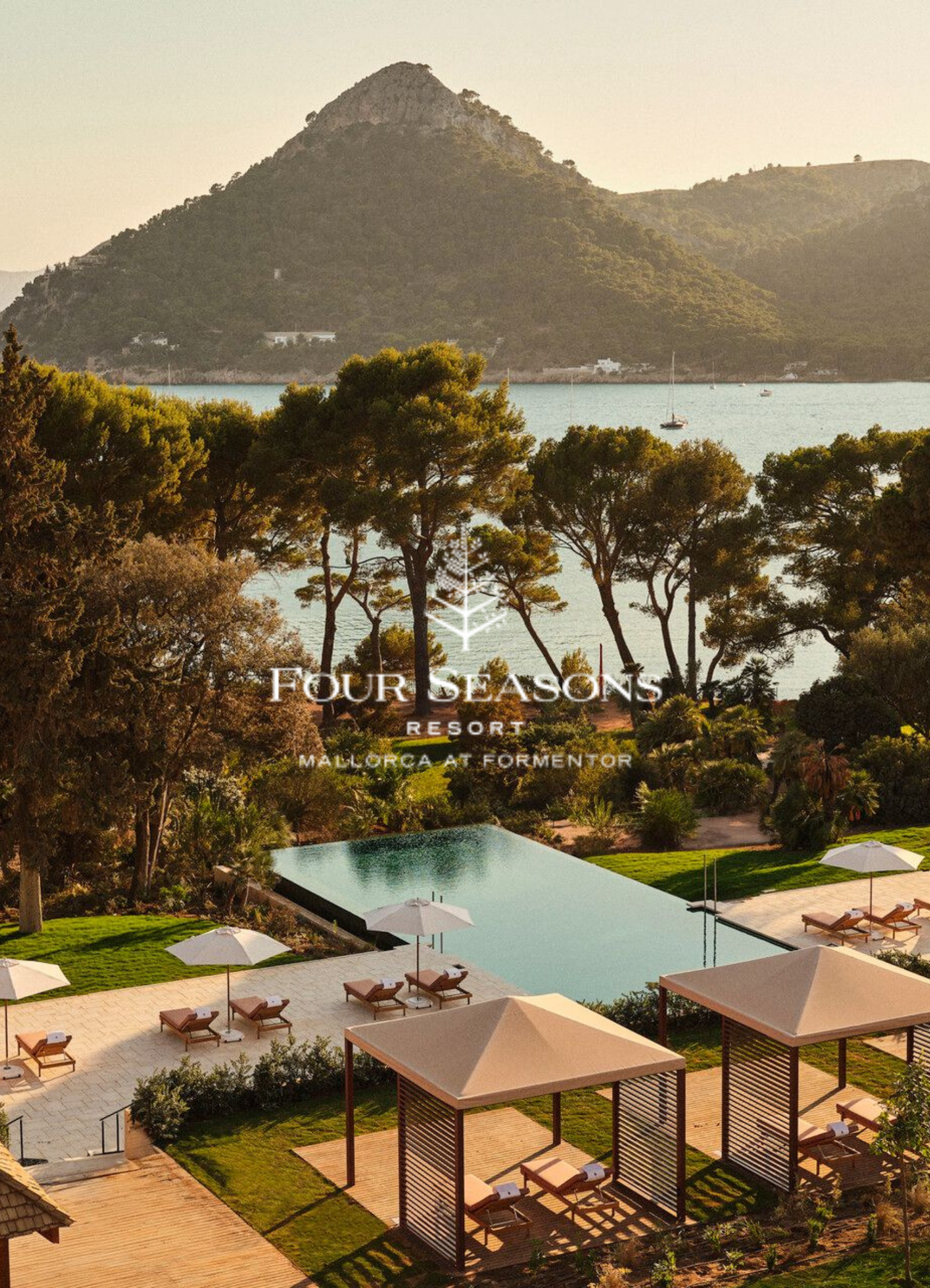 A view of a resort swimming pool area with lounge chairs and white umbrellas, surrounded by greenery, with a backdrop of mountains and a body of water with sailboats.