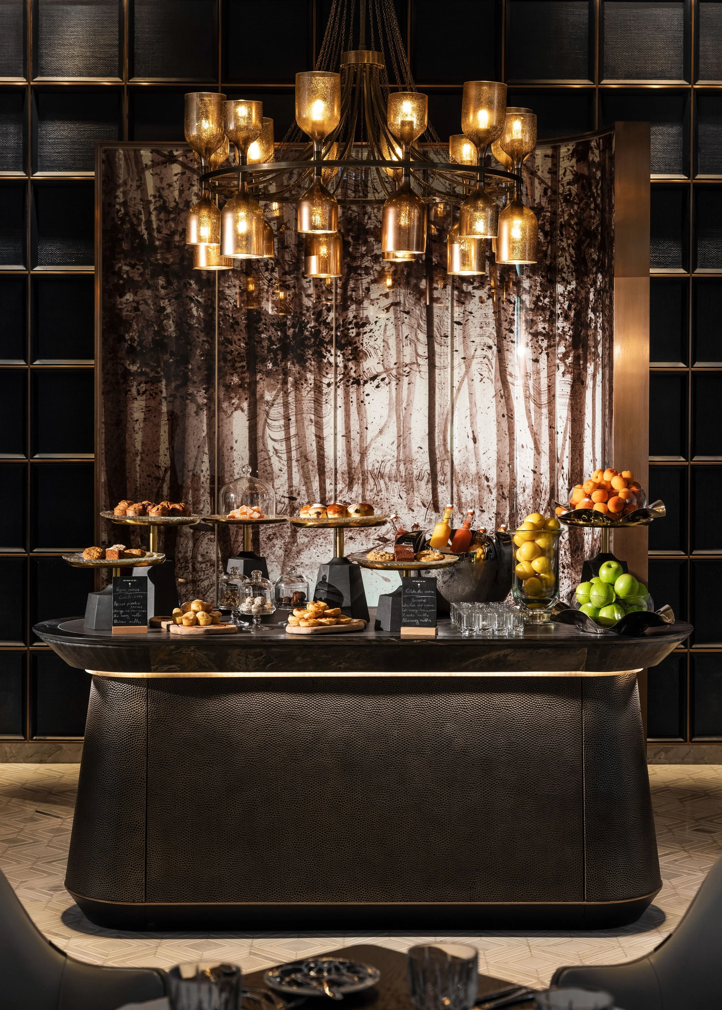 Elegant buffet table with an assortment of pastries, fruits, and beverages in a luxurious setting with a modern black and gold design, a large chandelier, and black shelving in the background.