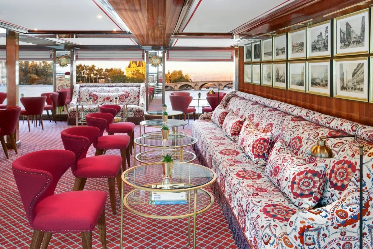 Interior of a stylish lounge or cafe with a long patterned sofa, red chairs, and glass tables, overlooking a river and historic bridge through large windows.