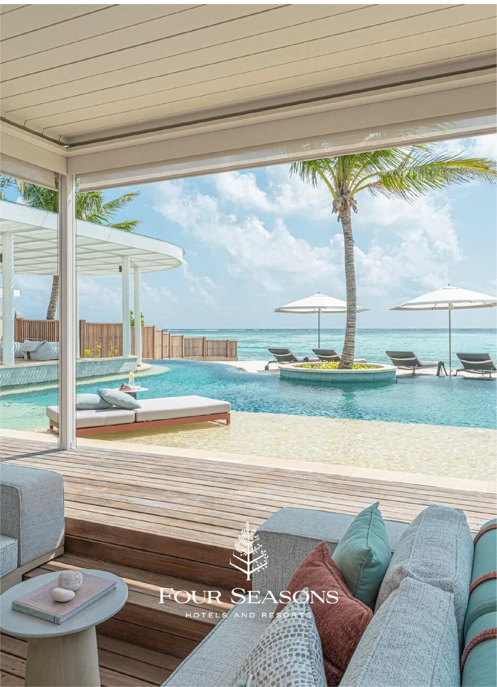 A view of a pool area at a beach resort with lounge chairs, umbrellas, palm trees, and the ocean in the background seen from a covered patio area with seating and pillows.