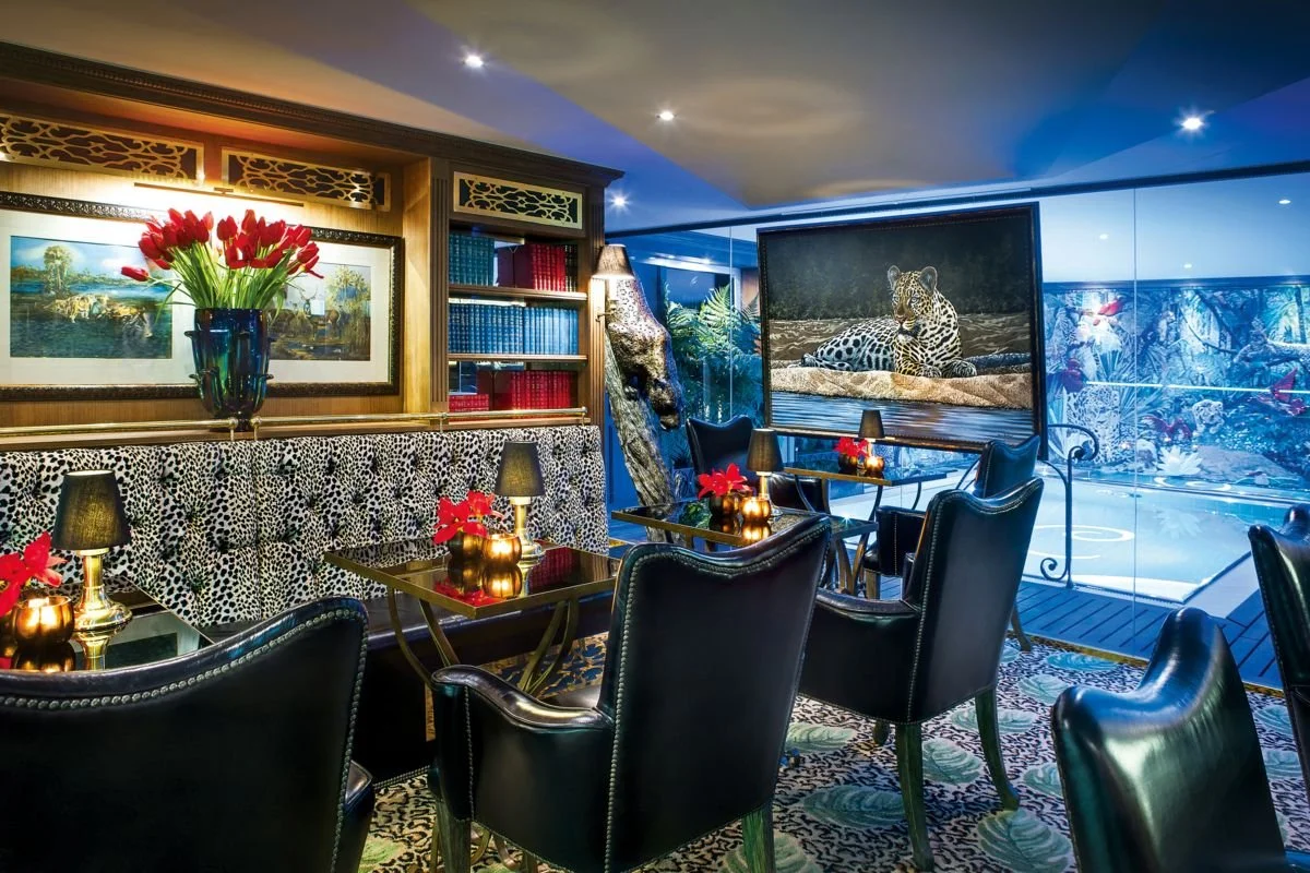 Luxurious lounge area with black leather chairs, small tables with red poinsettia flowers, a large vase of red tulips, a bookshelf with red and blue books, leopard-themed decor, a large framed painting, and a wildlife photograph of a leopard, with a glass wall showing an ice skating rink outside.