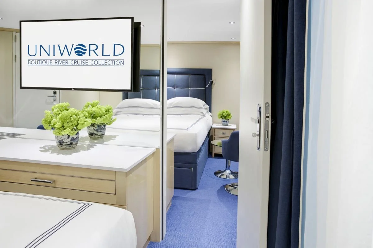 Hotel room with a bed, a nightstand with a plant, and a TV screen displaying 'UNIWORLD BOUTIQUE RIVER CRUISE COLLECTION'