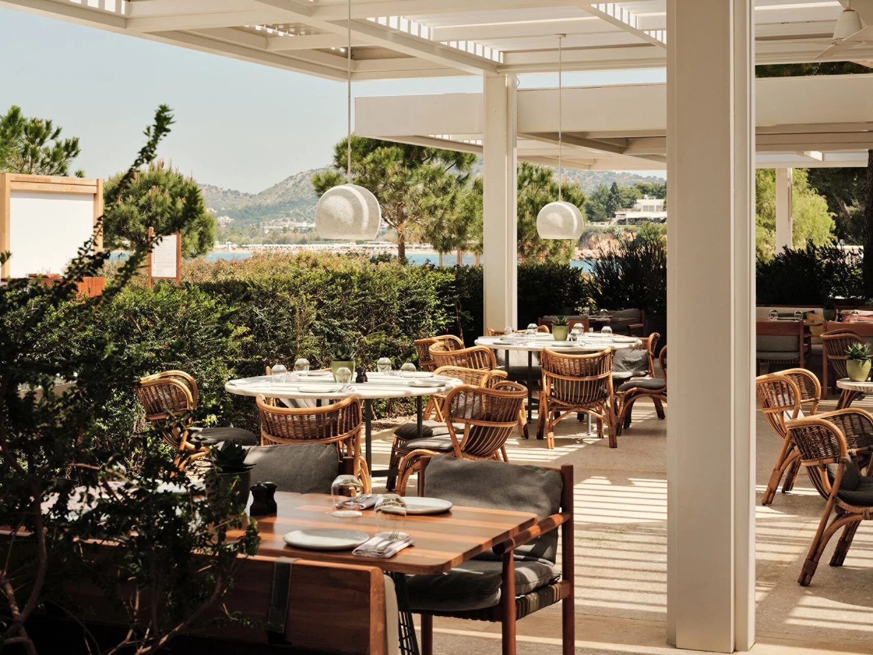 Outdoor restaurant patio with round tables set with plates and glasses, surrounded by wicker chairs, overlooking greenery and a body of water in the distance under a white canopy.