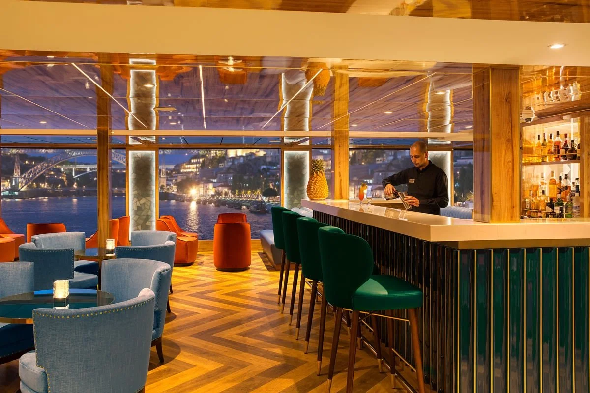 Elegant bar with green and blue chairs, a bartender, and a cityscape view of a river at dusk