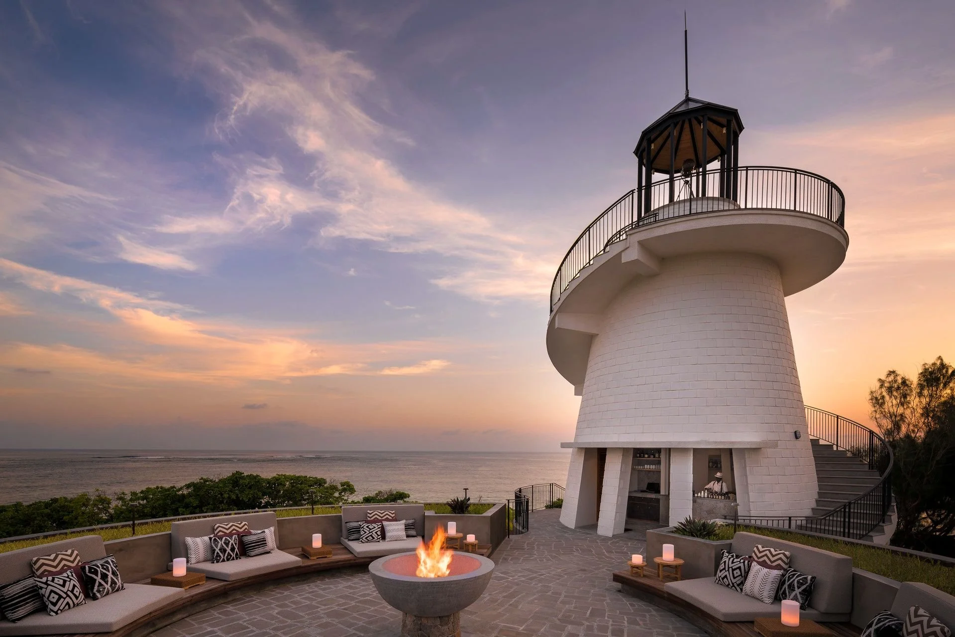 A white lighthouse with a black railing and a small enclosed observation area on top, situated near water during sunset. In the foreground, there is an outdoor seating area with couches, cushions, and a fire pit, creating a cozy atmosphere.