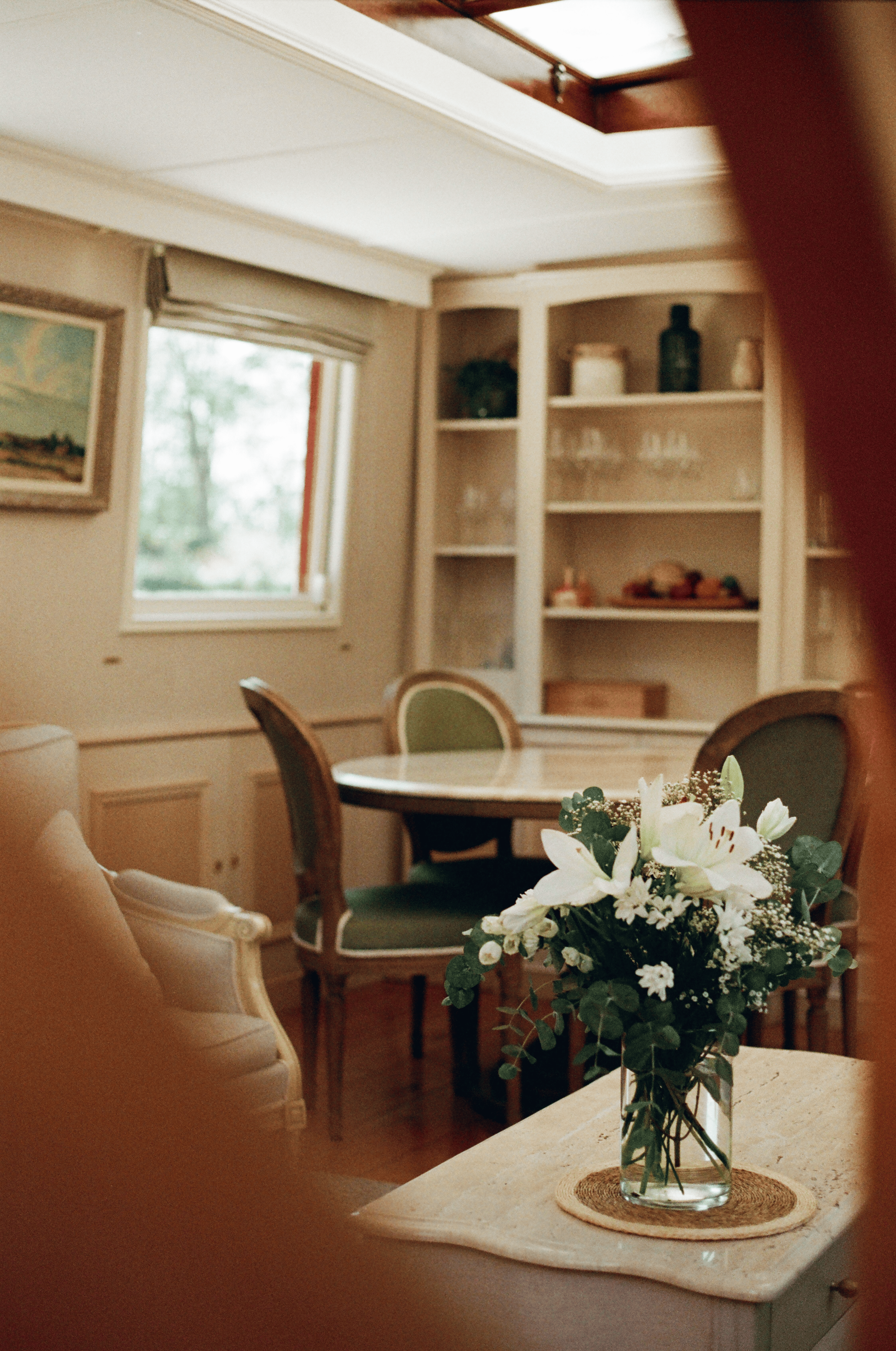 A living room with a white marble countertop, a bouquet of white lilies and other flowers in a glass vase on a woven placemat, and green upholstered chairs around a wooden dining table. There are built-in white shelves with decorative items and a window with a view of trees outside.