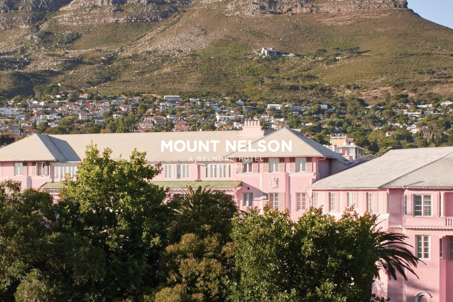 A mountain with rocky cliffs behind a suburban area with pink buildings and trees.
