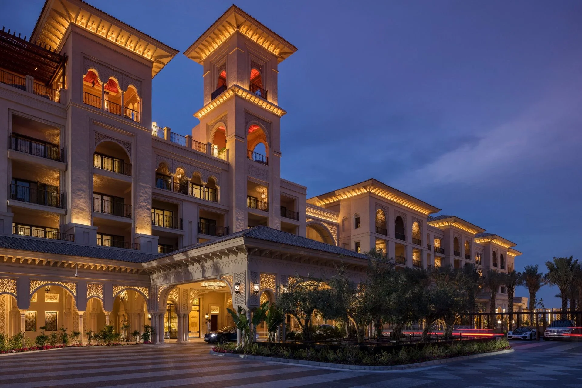 A multi-story luxury hotel with Mediterranean architecture, illuminated at dusk. The building features arches, balconies, and towers with warm lighting, set against a darkening sky. There are palm trees and a driveway with parked cars in the foreground.