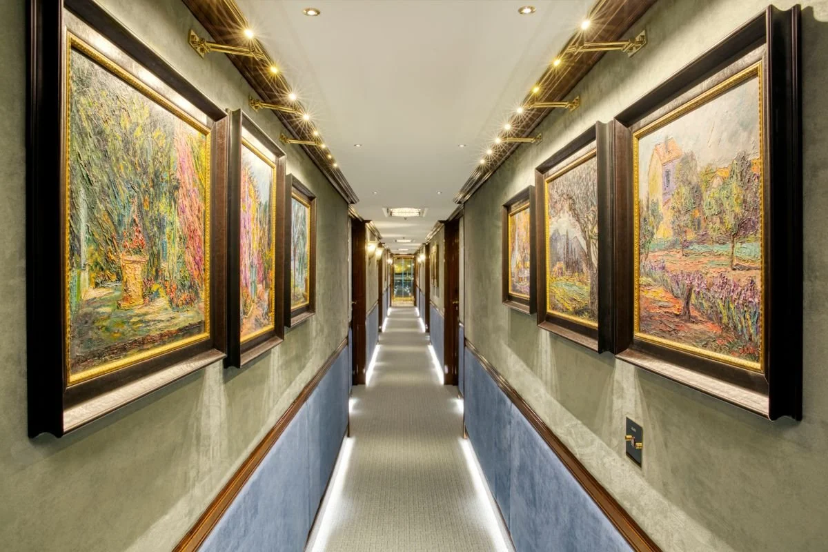 A hotel hallway with landscape paintings framed in gold, walls decorated with green and blue textured wallpaper, and recessed lighting on the ceiling.