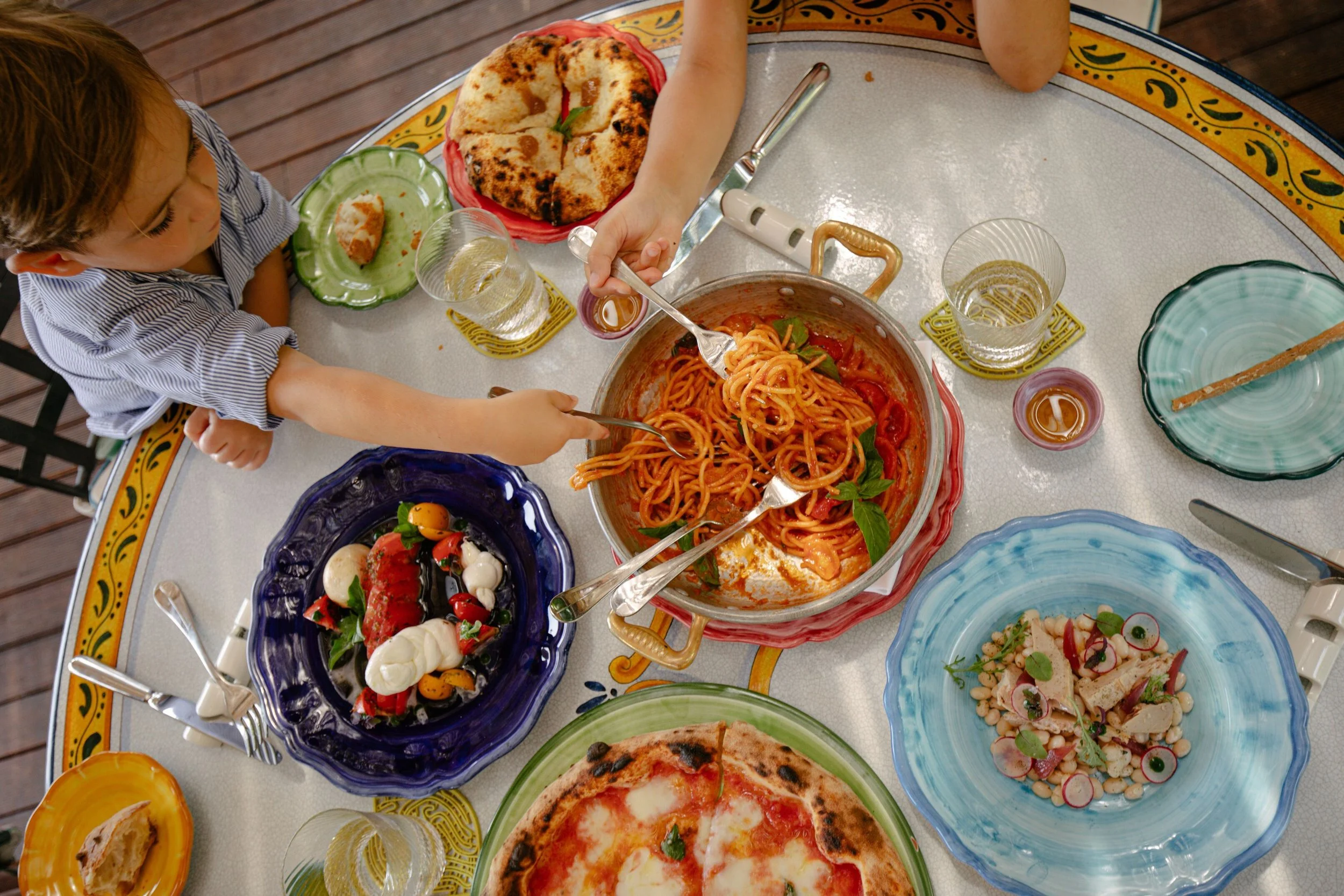 A top-down view of a dinner table with various colorful plates of food, including pasta, pizza, salads, and drinks, with two children reaching for the food.