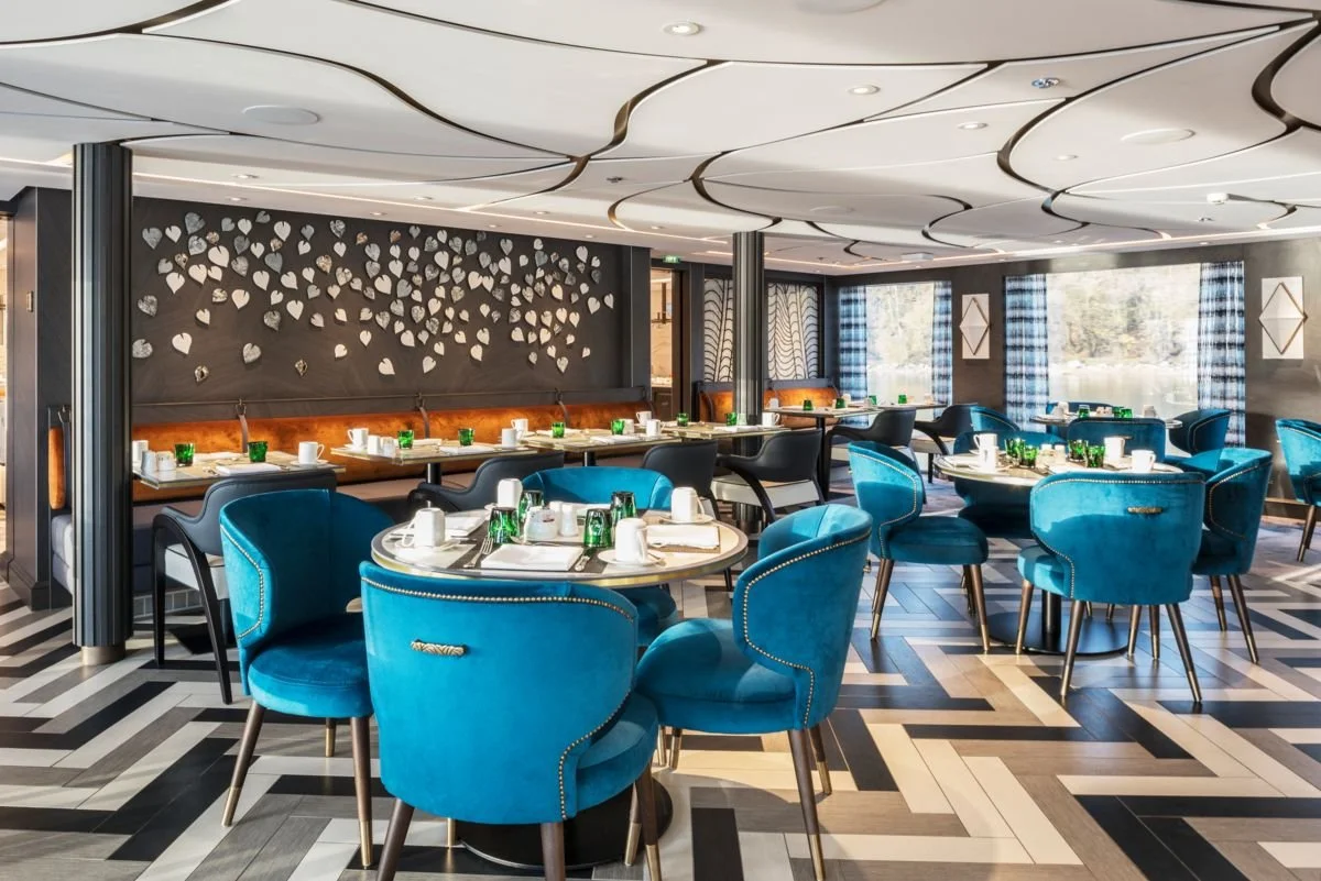 Elegant restaurant dining area with blue velvet chairs, round tables set with white cups and green glasses, and a wall decorated with 3D heart-shaped art. Large windows with blue plaid curtains let in natural light, and the ceiling features a modern, wavy design.