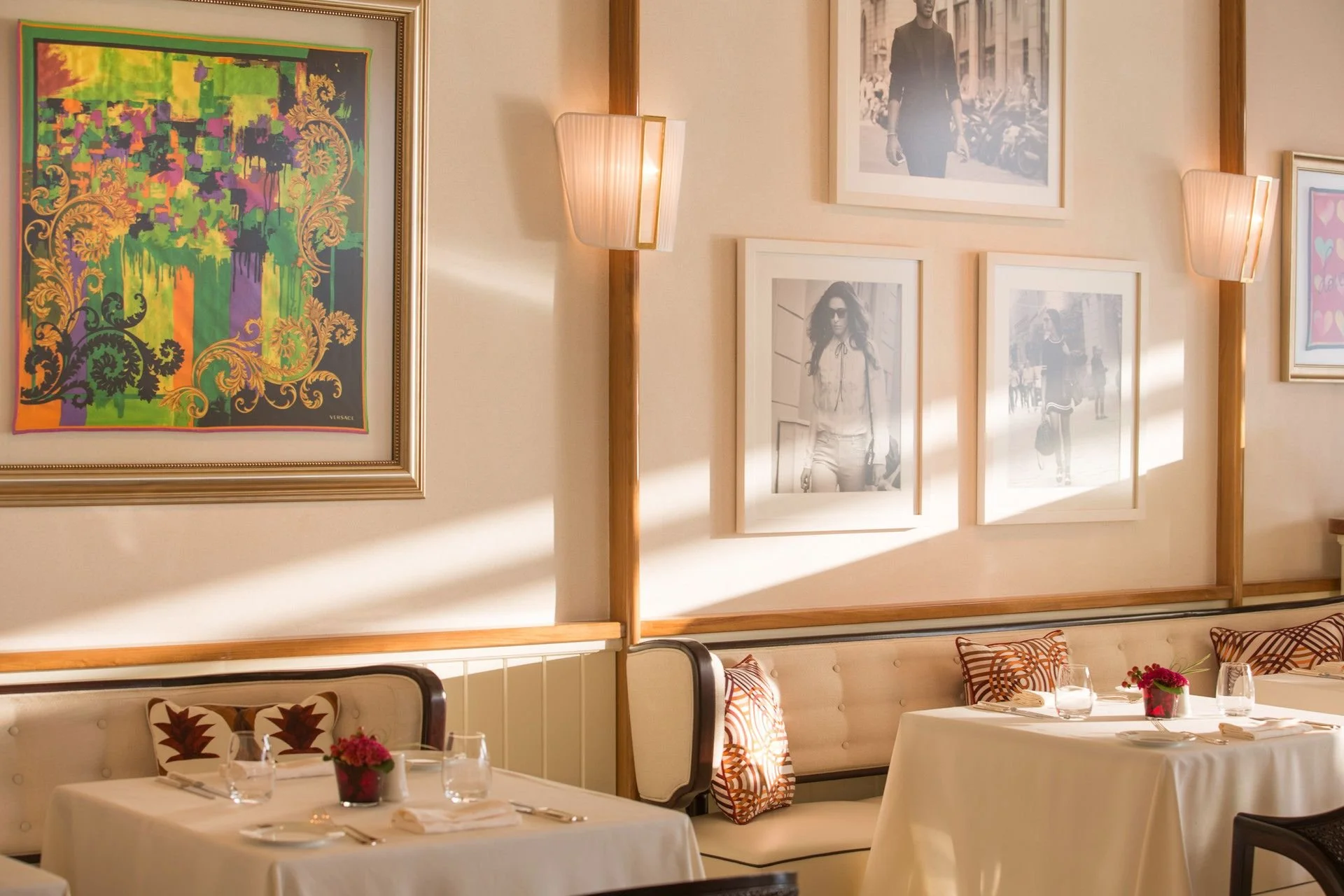 Elegantly set dining tables in a restaurant with floral centerpieces, white tablecloths, and framed black-and-white and colorful artwork on the walls, illuminated by wall sconces.