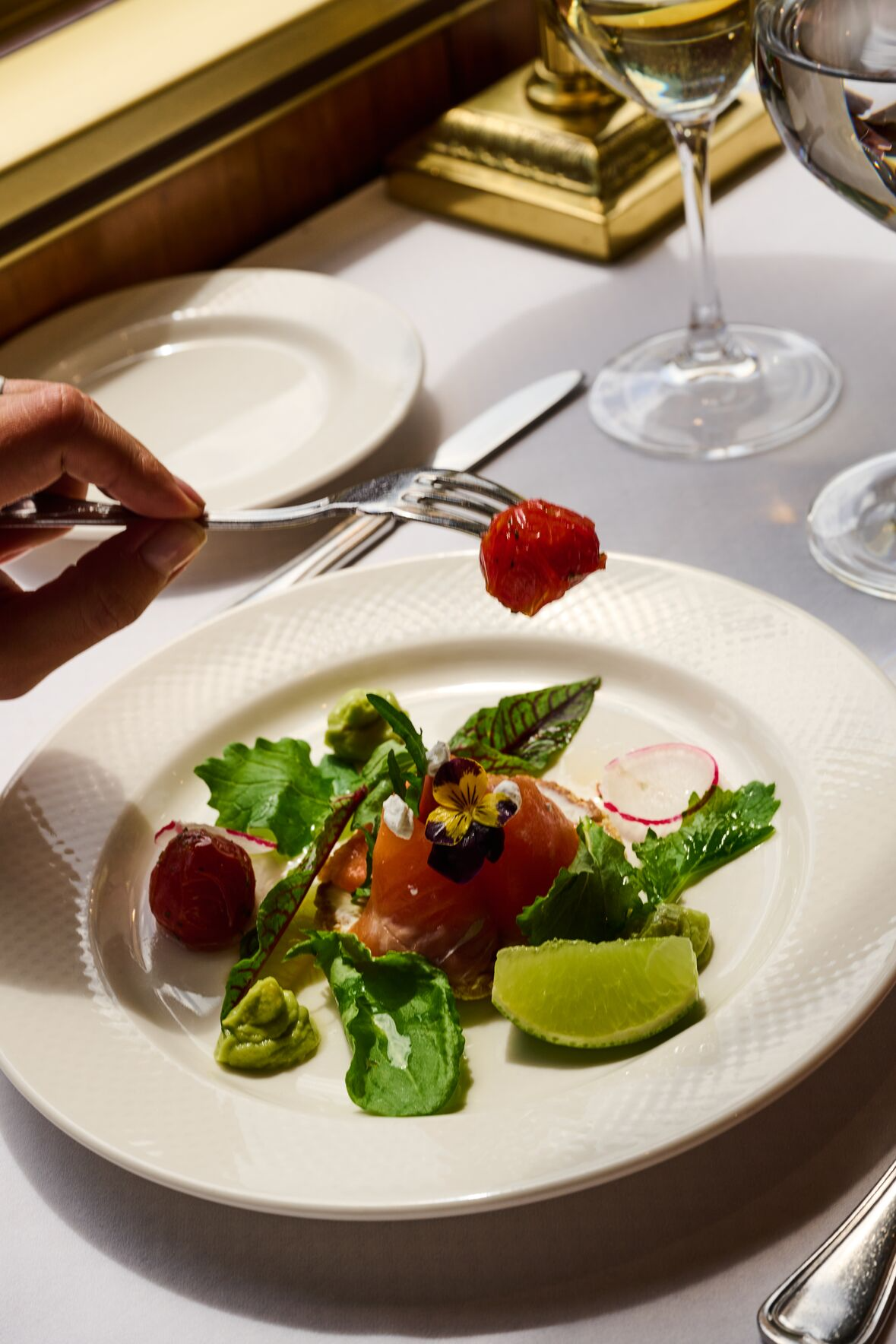 A hand holding a fork with a piece of tomato over a plated dish featuring greens, slices of avocado, tomato, a lime wedge, and decorative edible flowers, in a fine dining setting with glasses and tableware.