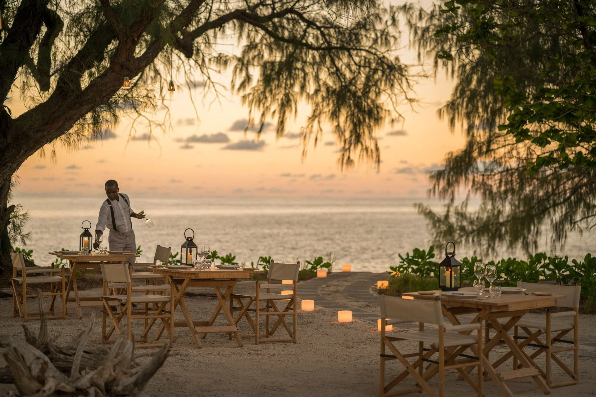 Beachside outdoor dining setup at sunset with tables, chairs, candles, and lanterns, overlooking the ocean, framed by trees.