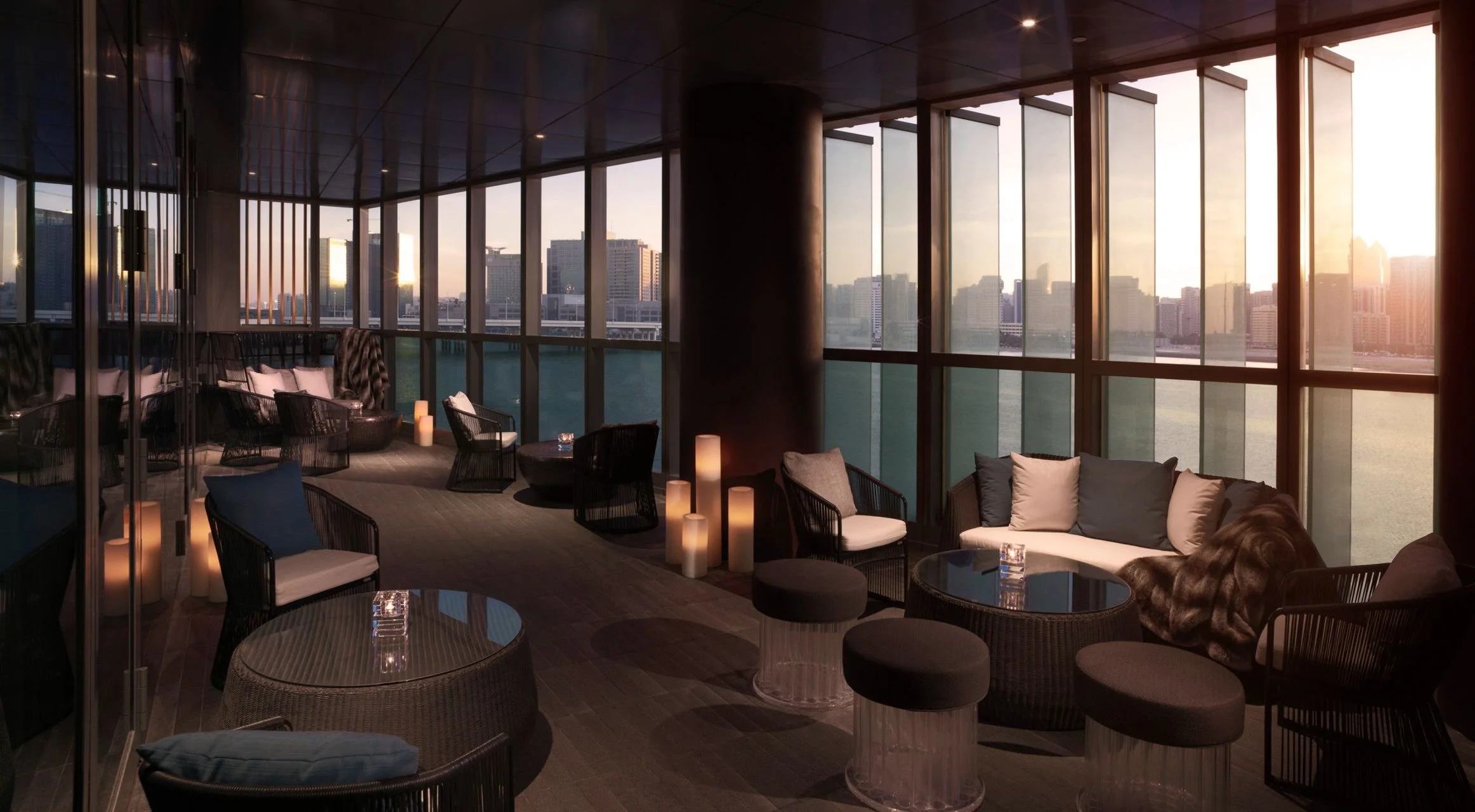 Interior of a high-rise lounge or apartment with large floor-to-ceiling windows showing an urban cityscape at sunset. The space has modern furniture including chairs, sofas, and round tables, with decorative candles and soft lighting creating a cozy ambiance.