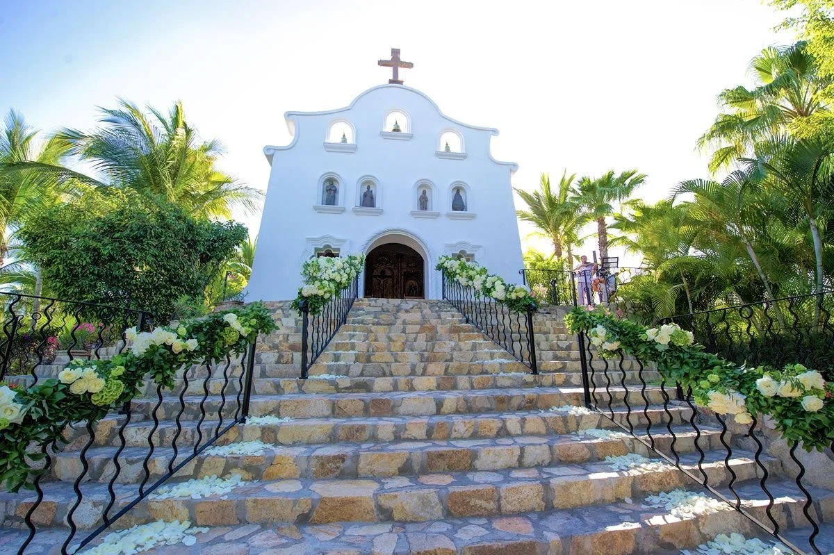 White chapel with a cross on top, floral decorations along the stairs, surrounded by green plants and palm trees, steps leading up to the entrance in a sunny outdoor setting.