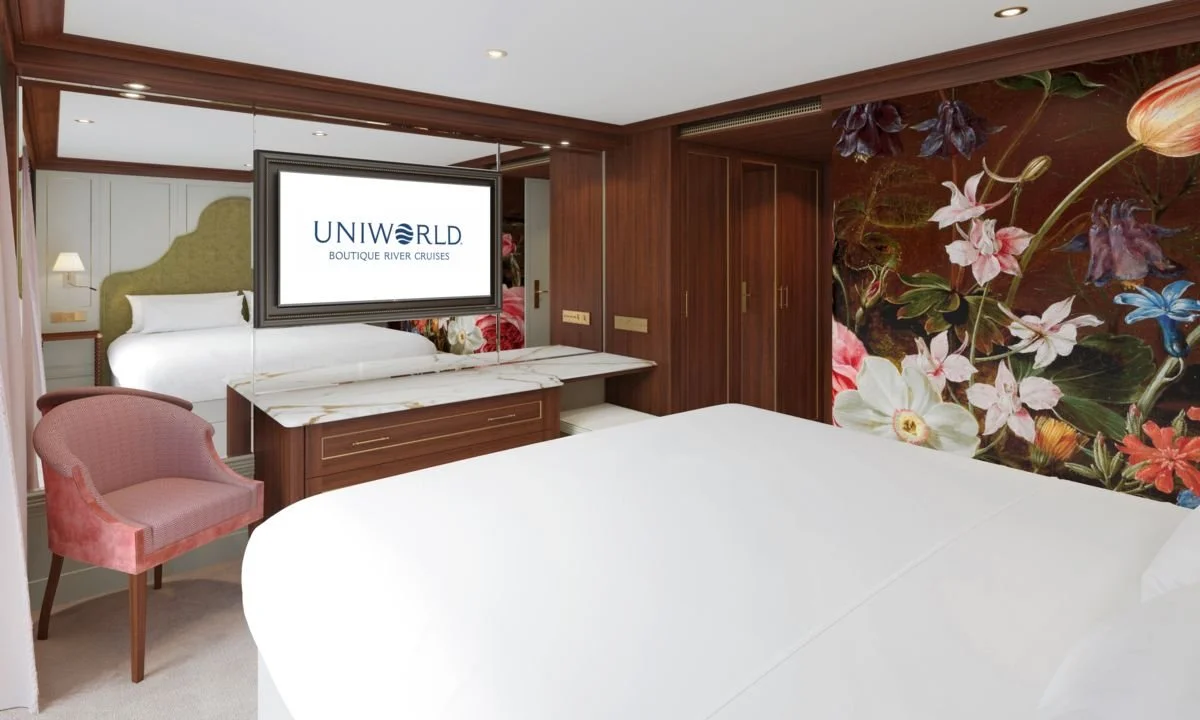 A hotel room with a floral accent wall, a large bed, a pink upholstered chair, and a mounted flat-screen TV displaying 'UNIWORLD BOUTIQUE RIVER CRUISES'.