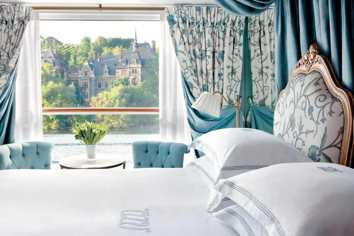 Luxurious hotel room with a large window overlooking a river and historic buildings, decorated with blue and white floral curtains, a bed with a tufted headboard, and a small round table with a vase of white tulips.