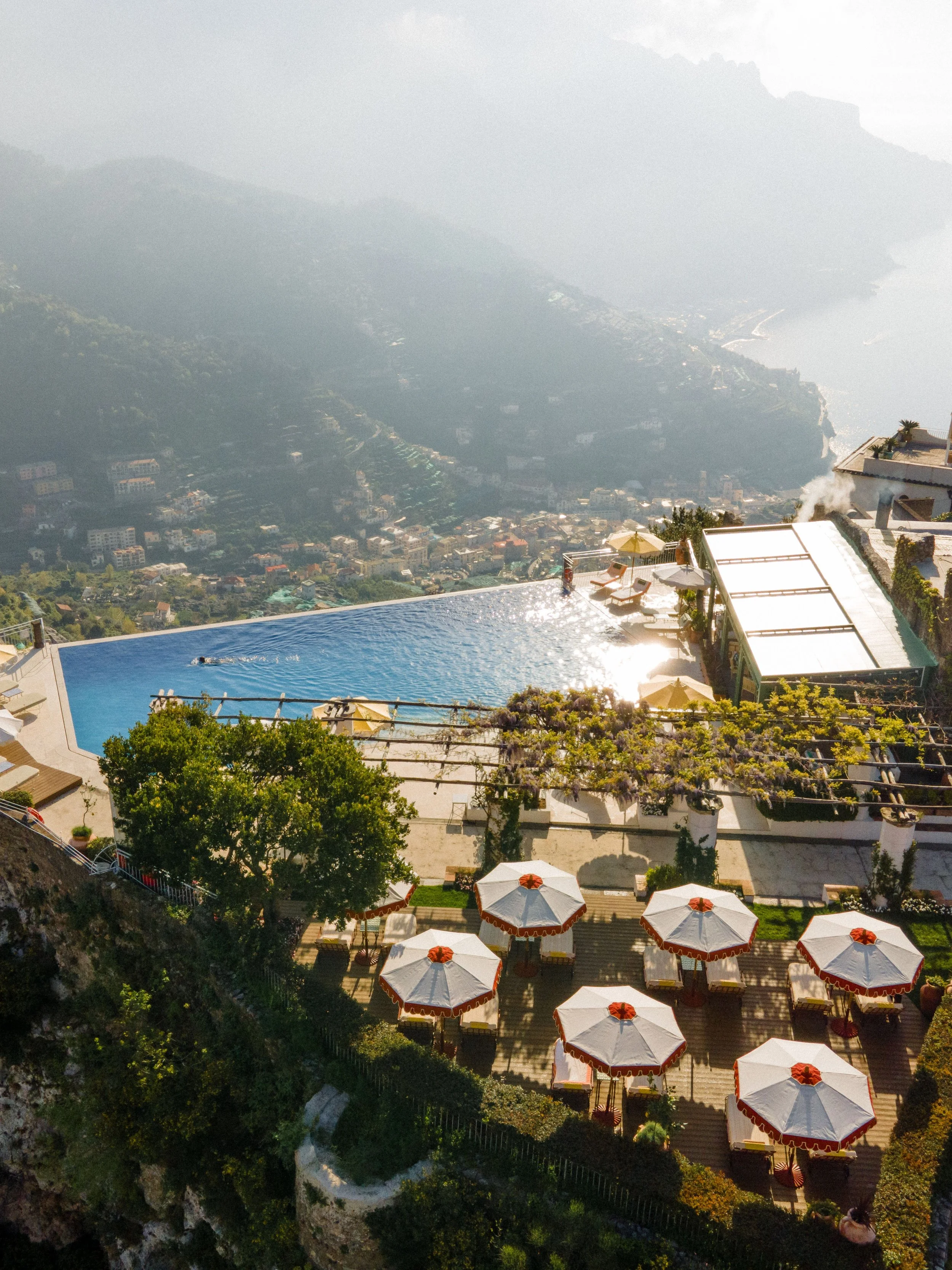 An outdoor infinity pool with umbrellas and loungers overlooking a cityscape and mountains in the distance.
