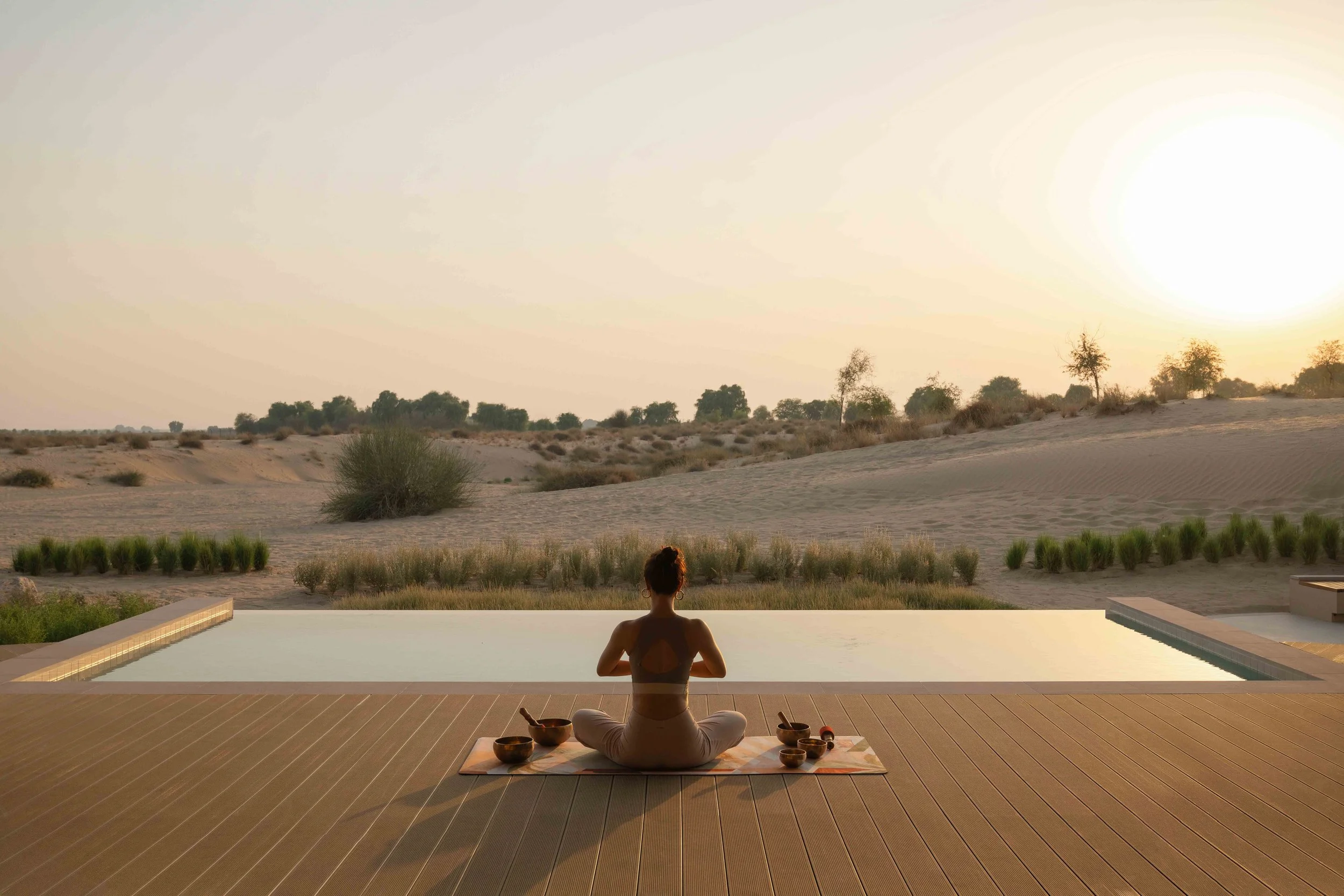 A woman practicing yoga and meditation outdoors on a wooden deck at sunset, with a desert landscape and sparse trees in the background.