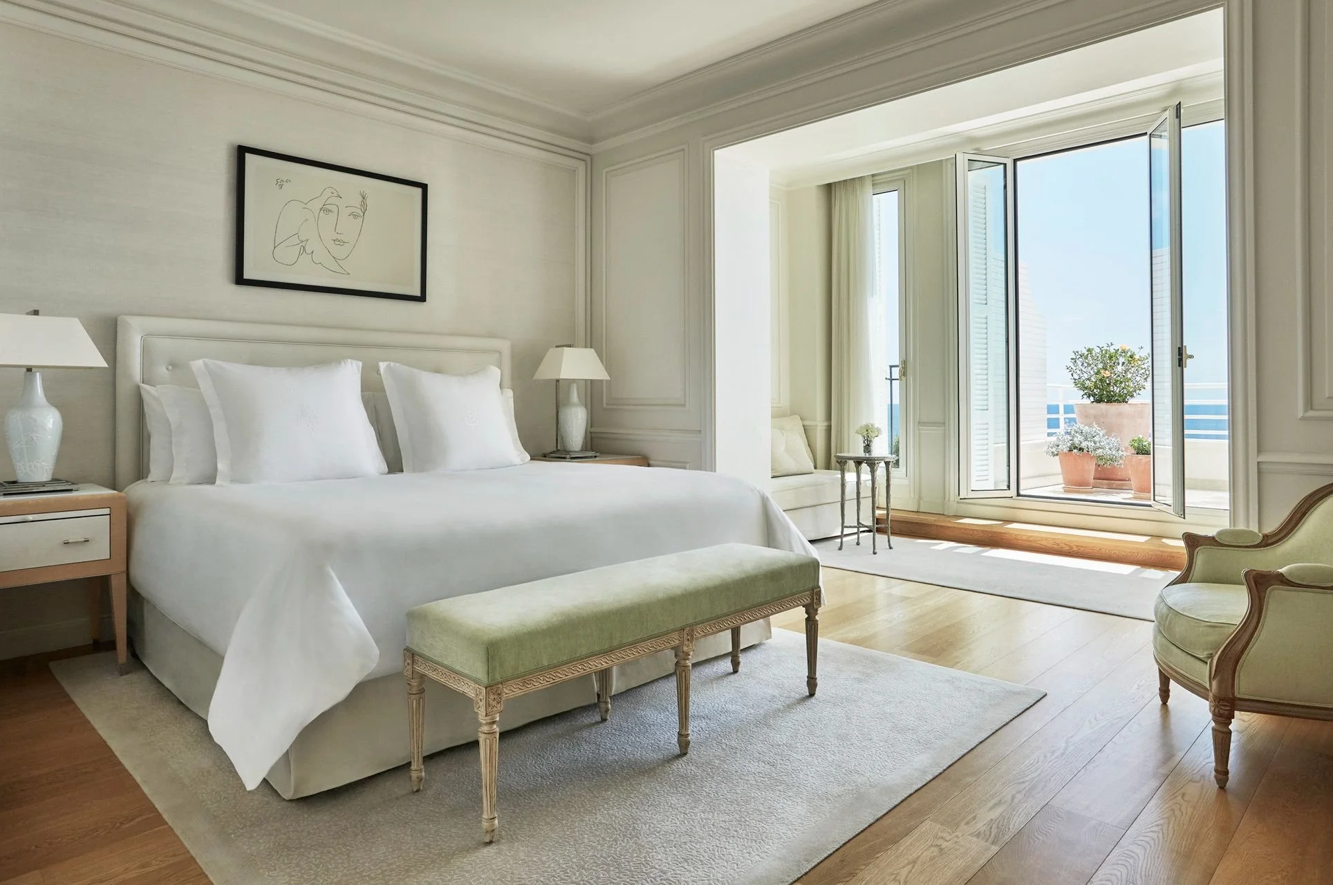 A bright hotel bedroom with a large bed, two bedside lamps, a framed abstract line drawing above the bed, and a balcony door with outdoor potted plants visible through the window.
