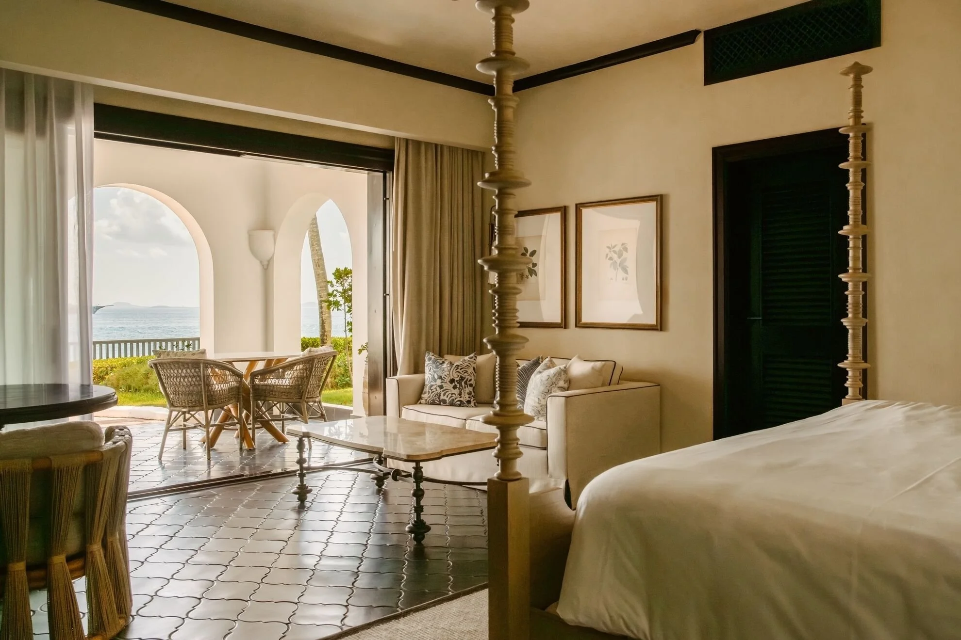 Hotel room with a living area and a view of the ocean through open sliding doors. The living area includes a beige sofa with pillows, a coffee table, and framed artwork on the wall. There is a small balcony with wicker chairs and a table outside.