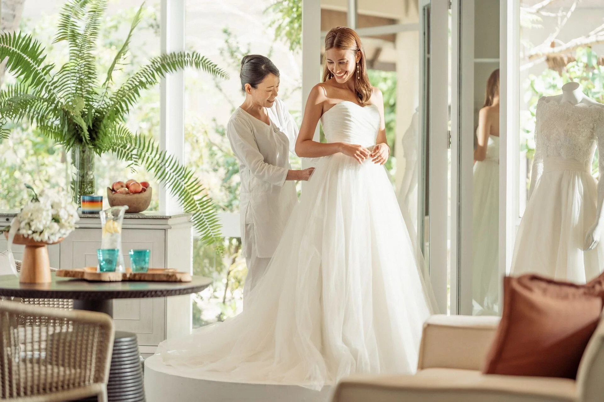 A bride in a white wedding dress standing on a platform in a bright room, smiling, while a woman assists her with the dress. There are mannequins with wedding dresses and lush green plants in the background.