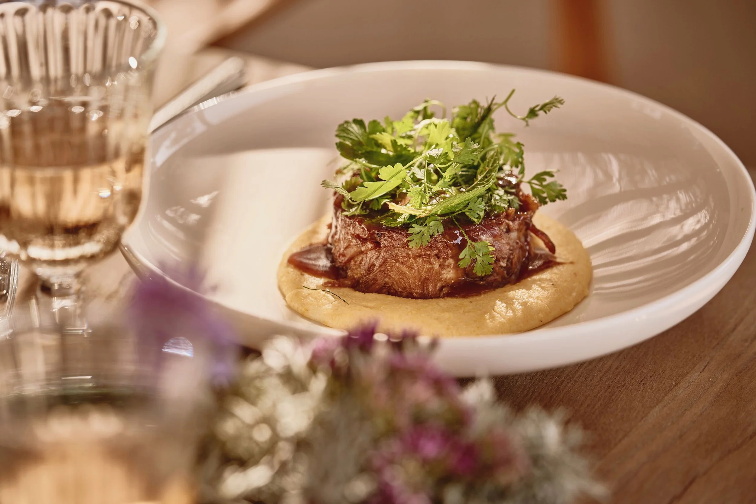A plated gourmet dish featuring tender meat garnished with fresh herbs on a bed of mashed potatoes or similar accompaniment, with a glass of water nearby.