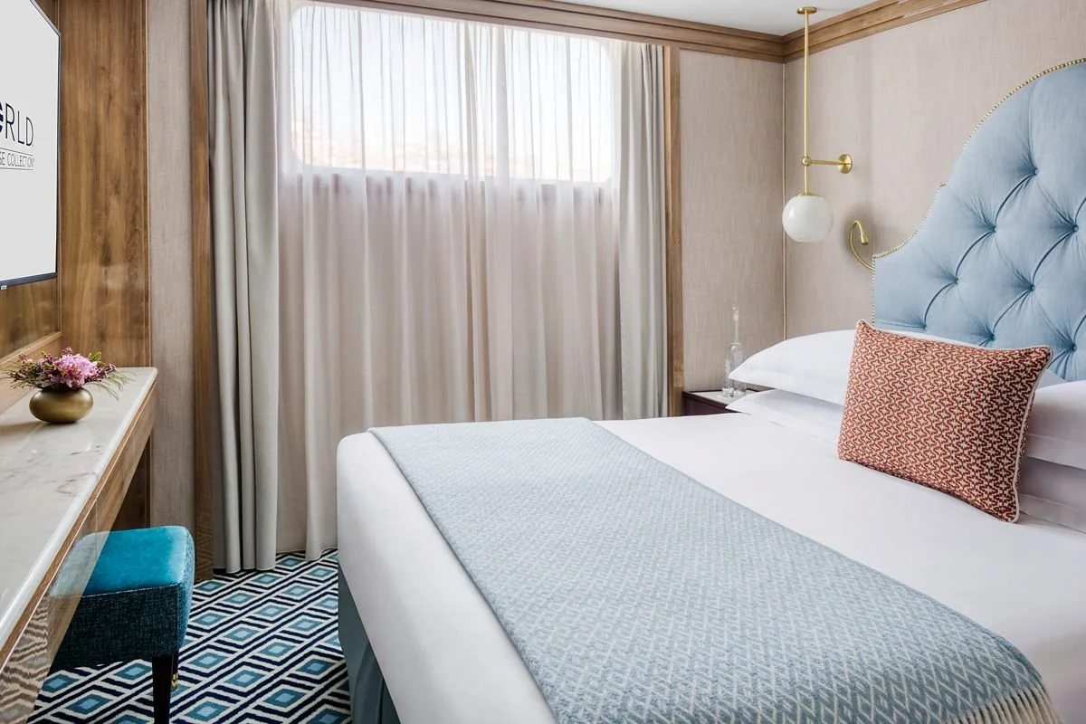 A neatly made hotel room bed with a light blue tufted headboard, white sheets, a red patterned pillow, and a light blue bed runner. There is a window with white curtains, a bedside table with a glass water bottle, a wall-mounted white sconce, and a corner with a blue velvet stool. A flat-screen TV is mounted on the wall to the left, with a small flower arrangement on a wooden shelf.